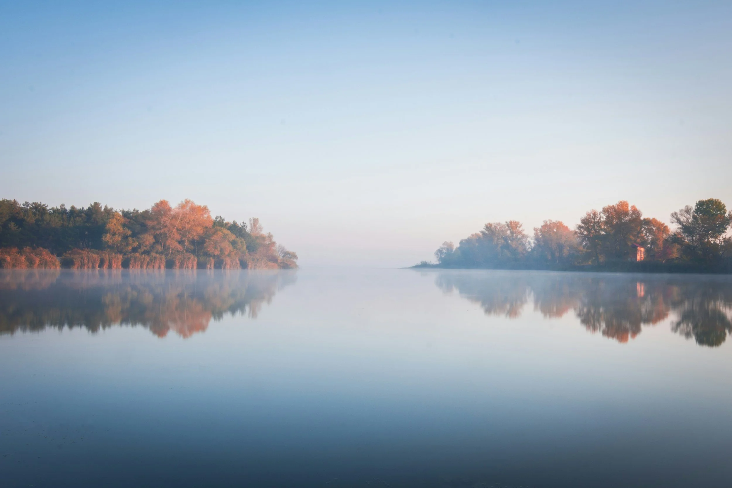 Calm river reflecting trees with orange and green foliage under a clear sky.