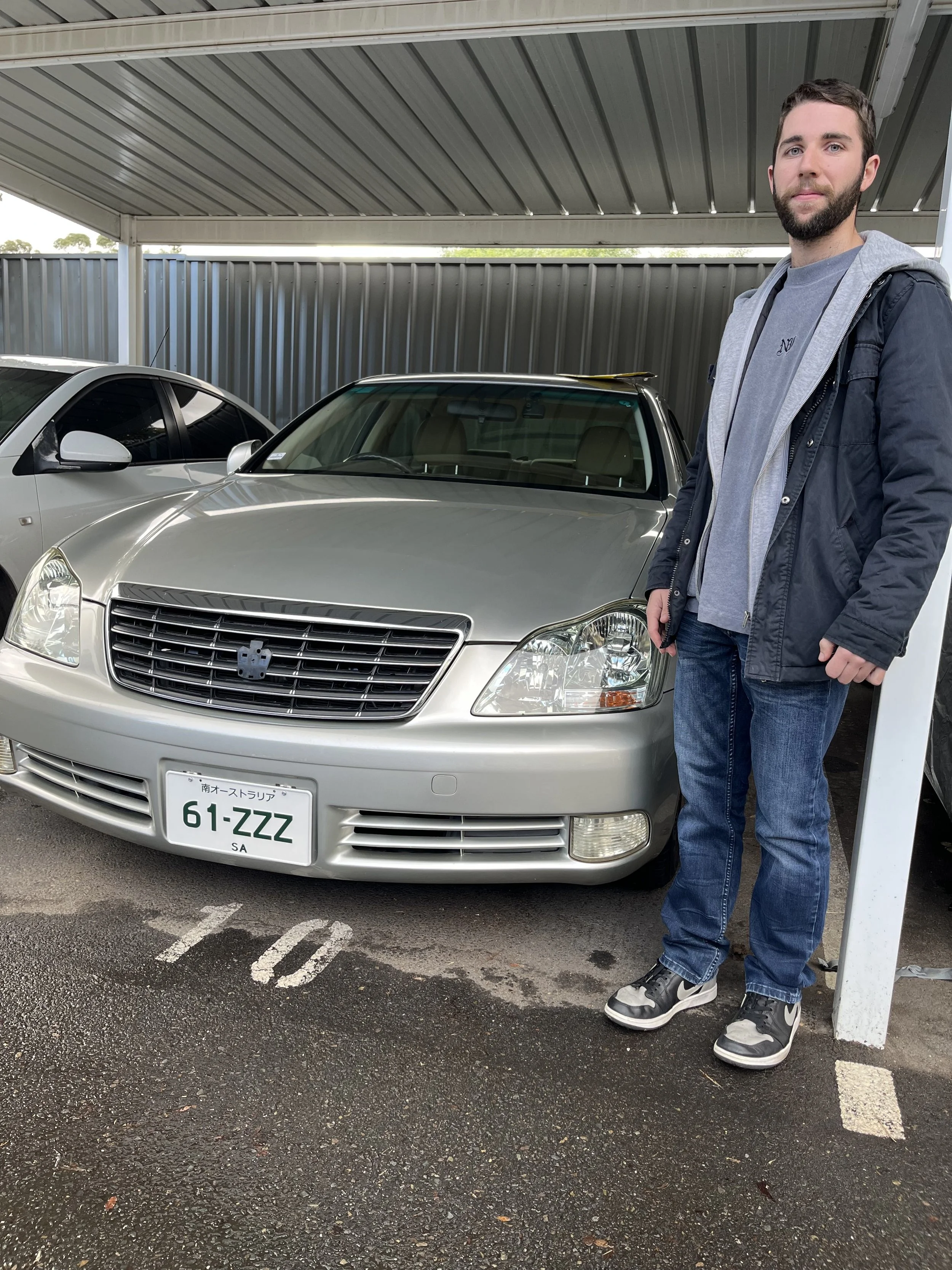 A man with a beard and blue eyes standing next to a silver luxury car in a parking lot under a metal roof, wearing a gray hoodie, dark jacket, jeans, and Nike sneakers.