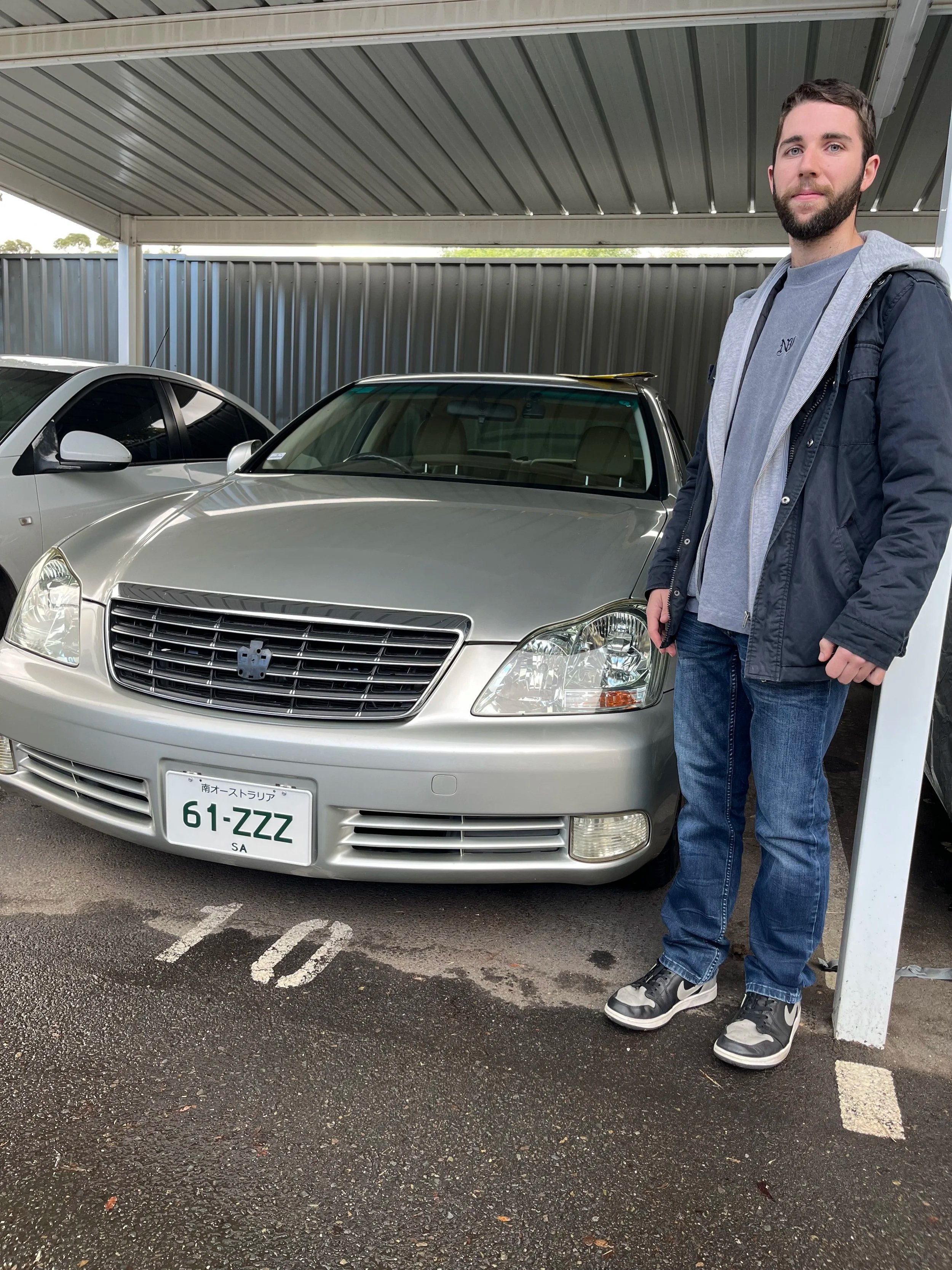 A man standing beside a silver car in a parking lot under a metal canopy.