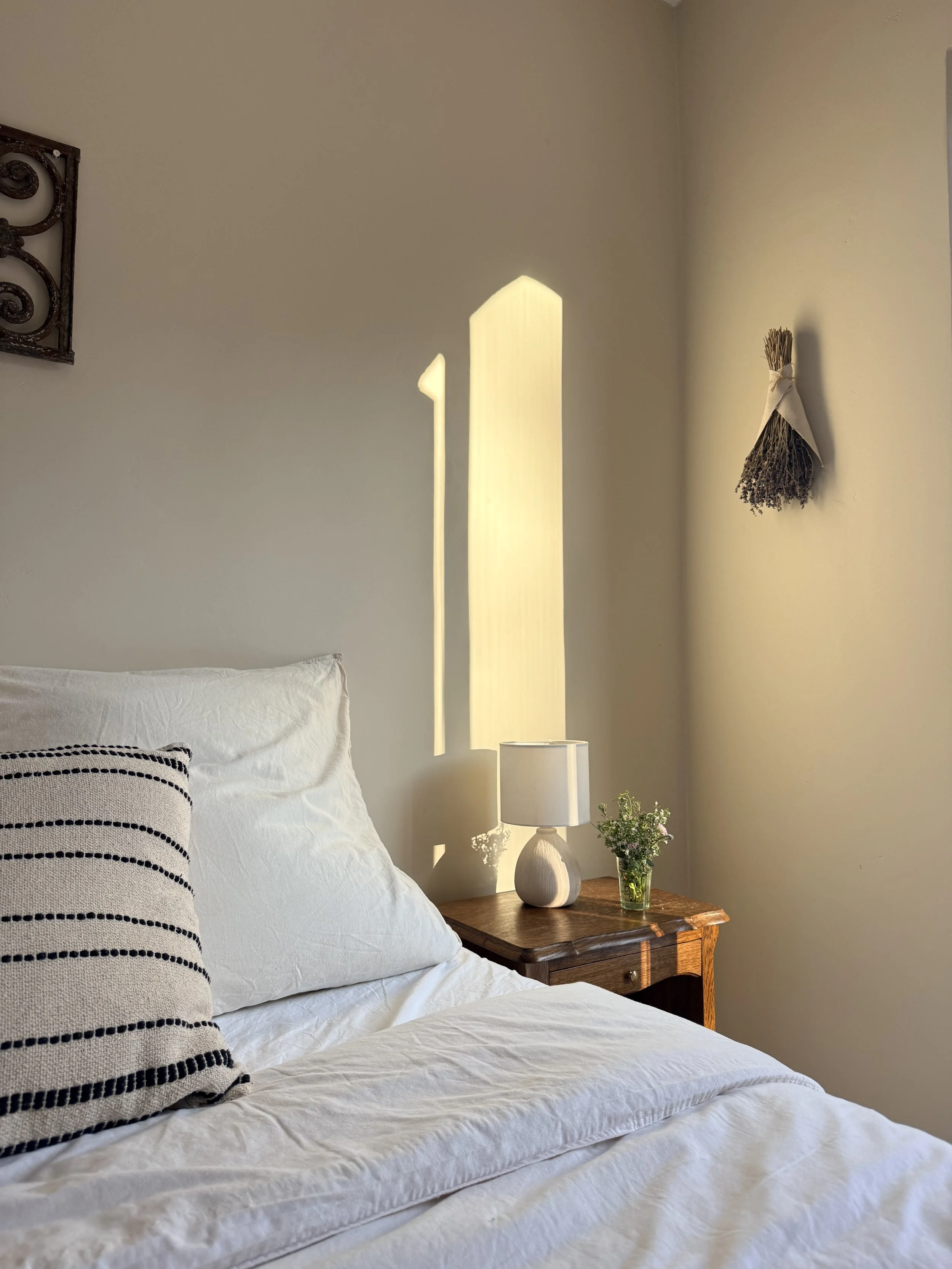 A cozy bedroom corner with a bed covered in white bedding, a black-and-white striped pillow, a wooden nightstand holding a white ceramic lamp, and a small vase of flowers. The wall has a hanging bundle of dried herbs tied with a ribbon and a decorati