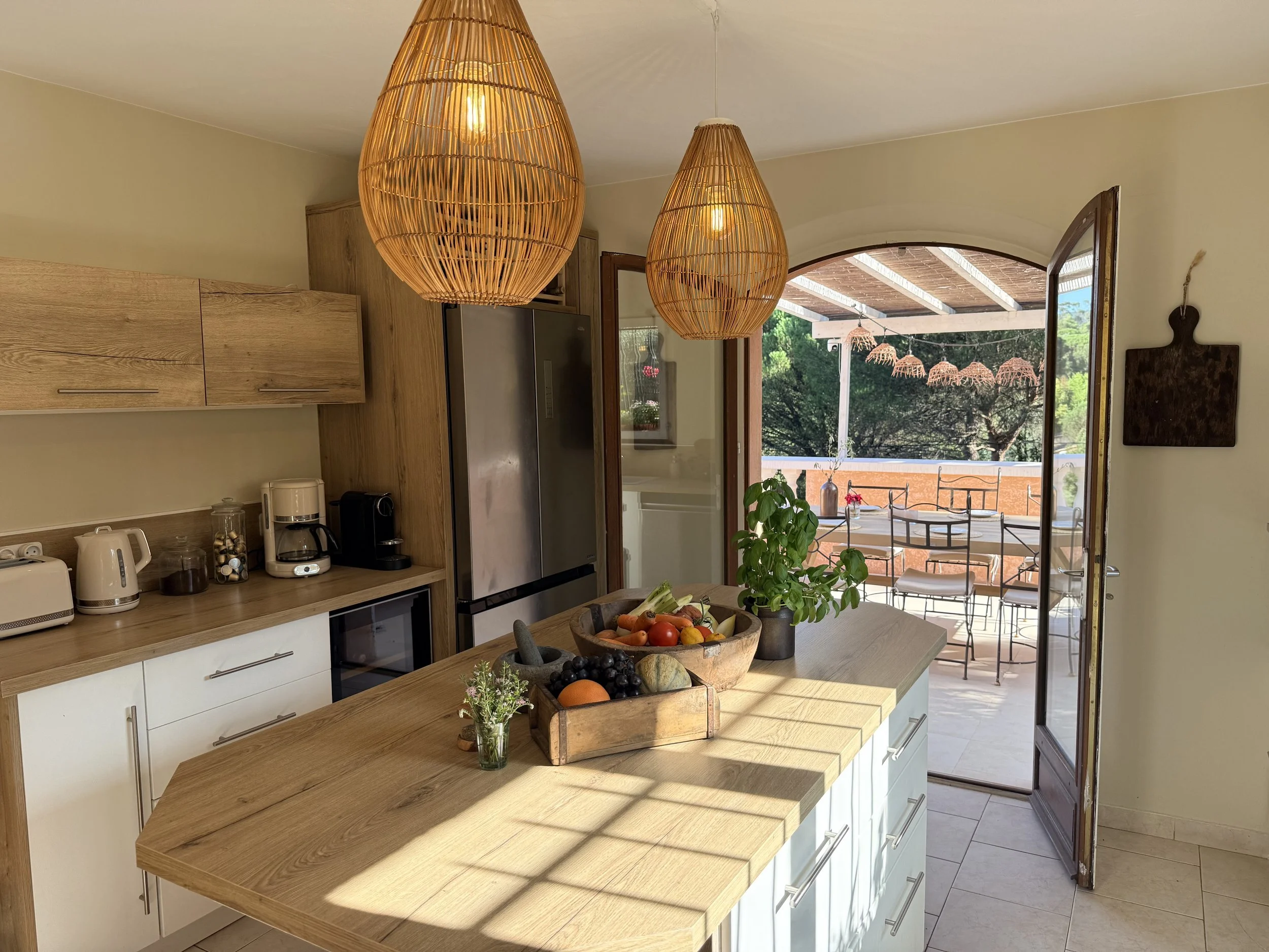 Kitchen with wooden countertops and hanging wicker pendant lights, open to outdoor patio with tables and chairs, and lush greenery in the background.