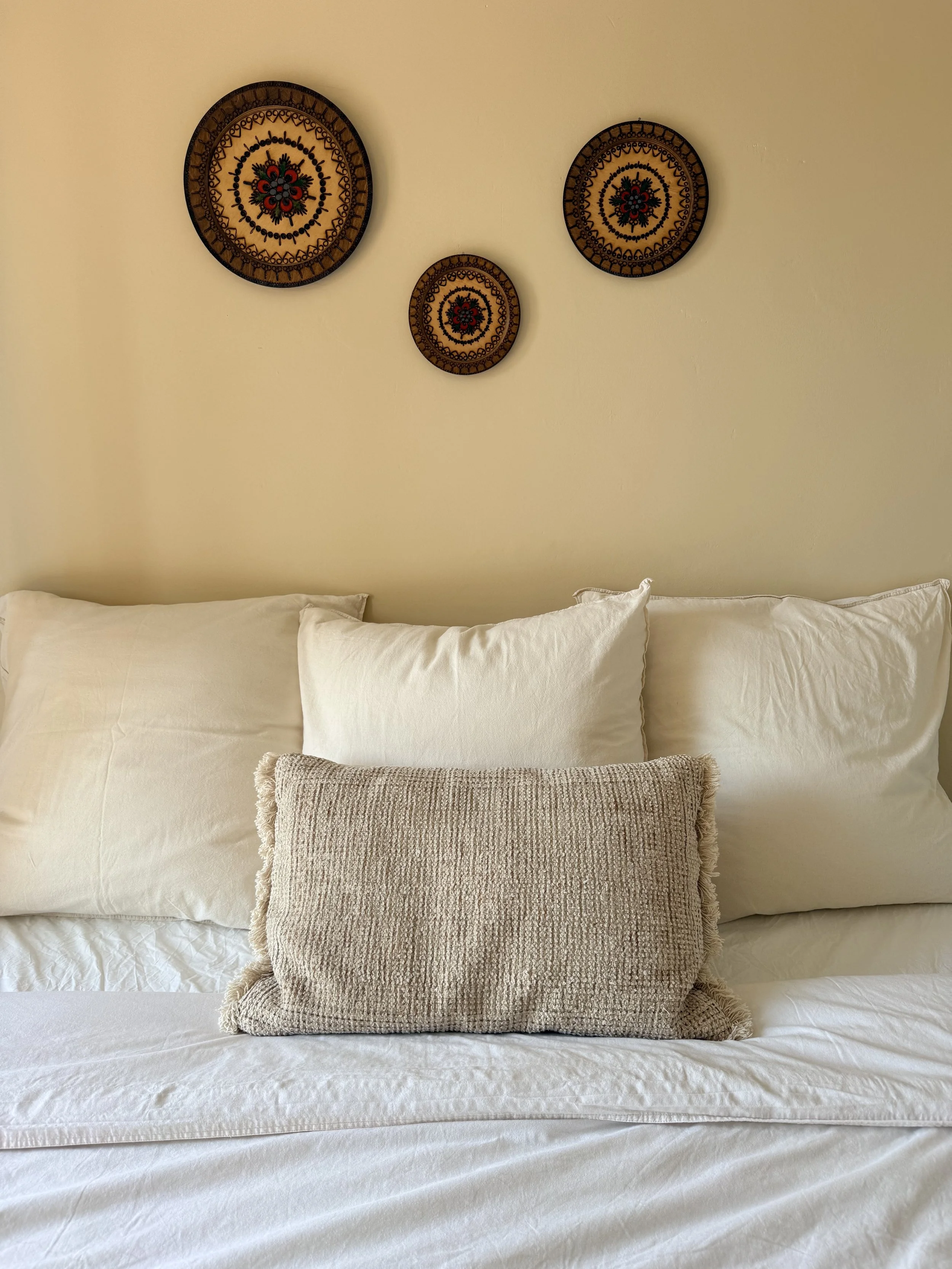 A neatly made bed with white sheets and pillows, topped with a textured beige throw pillow, and three decorative circular wall hangings above the bed on a beige wall.
