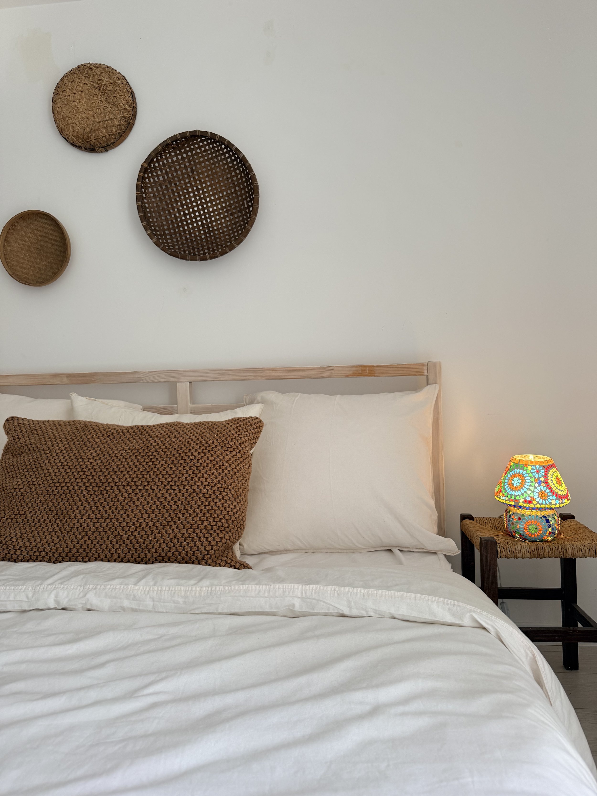 A neatly made bed with white linens and a large brown textured pillow, next to a wooden nightstand with a colorful mosaic lamp, and a light green wall decorated with three woven baskets.