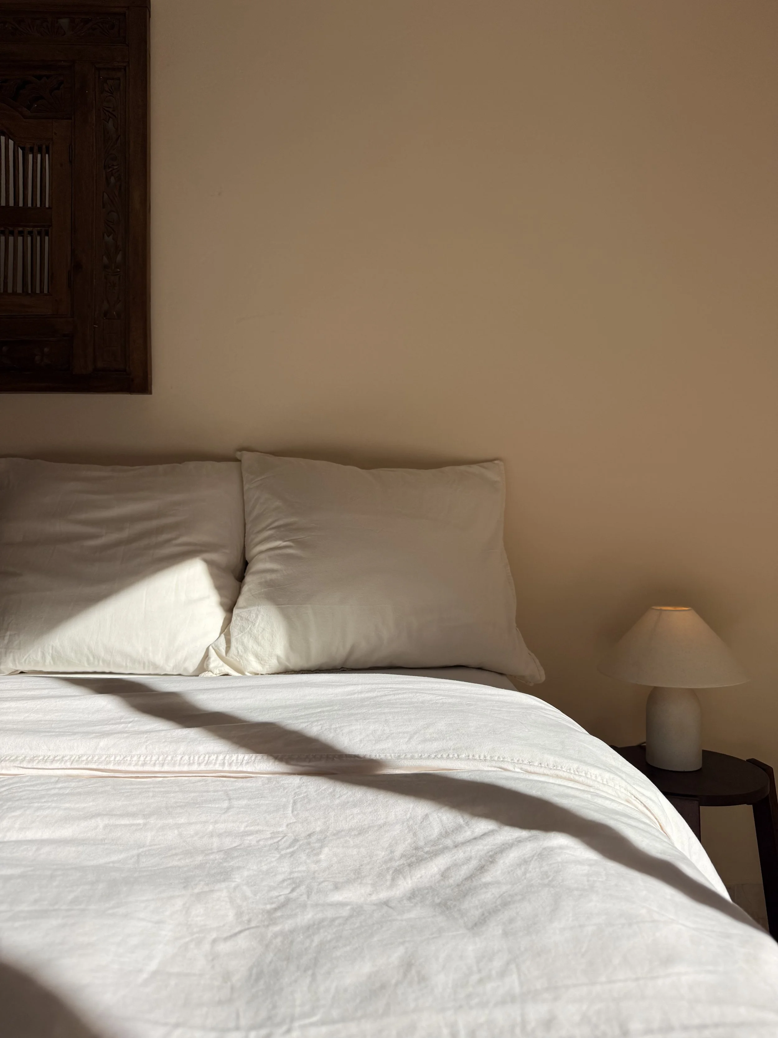 A neatly made bed with white sheets, two white pillows, a wooden headboard, a small bedside table with a lamp, on a beige wall.