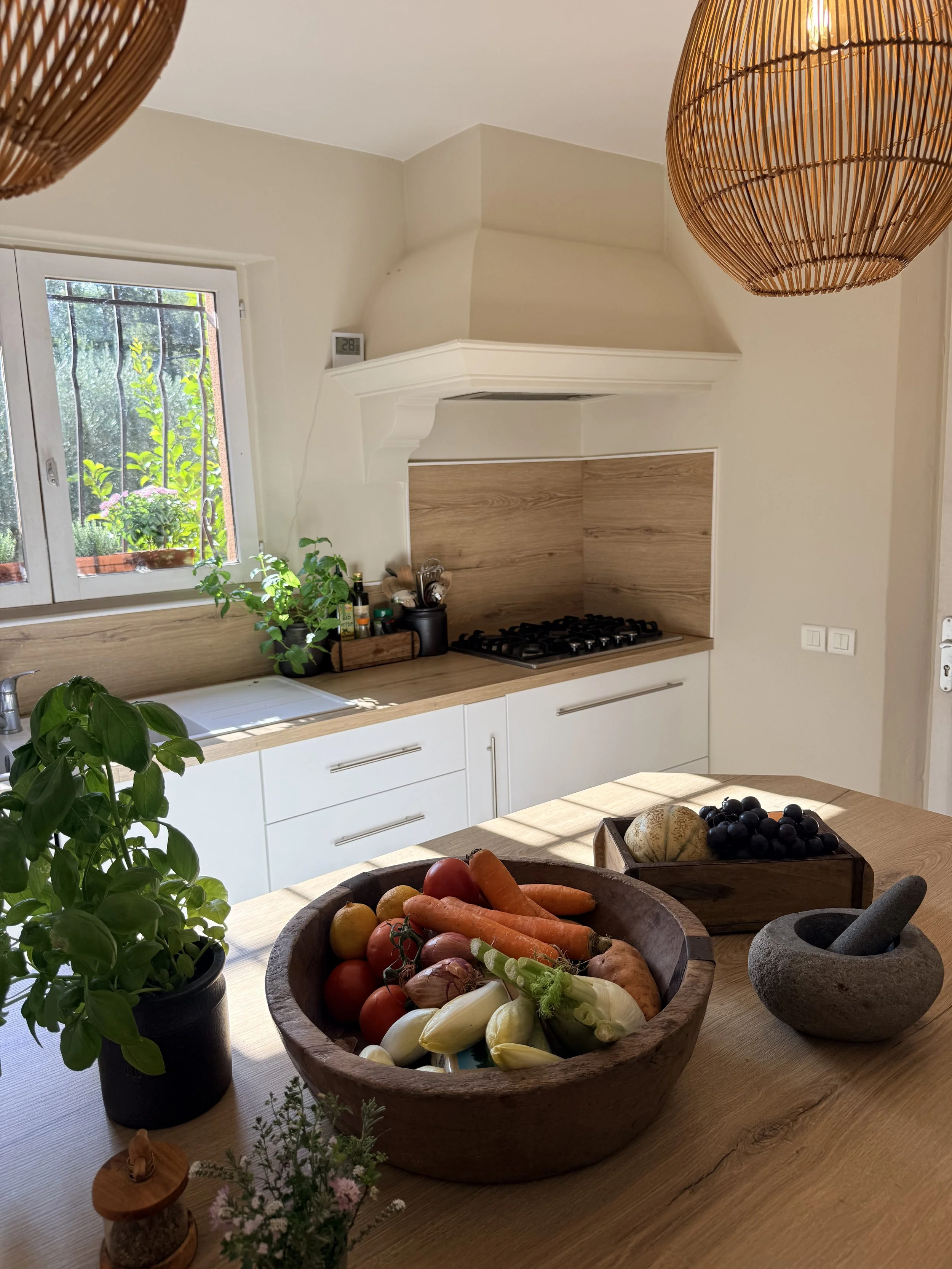 A kitchen with a window, plants, and a wooden dining table. On the table, there is a large wooden bowl filled with vegetables including tomatoes, carrots, onions, and garlic. There are also decorative stones and small plants on the table. The kitchen
