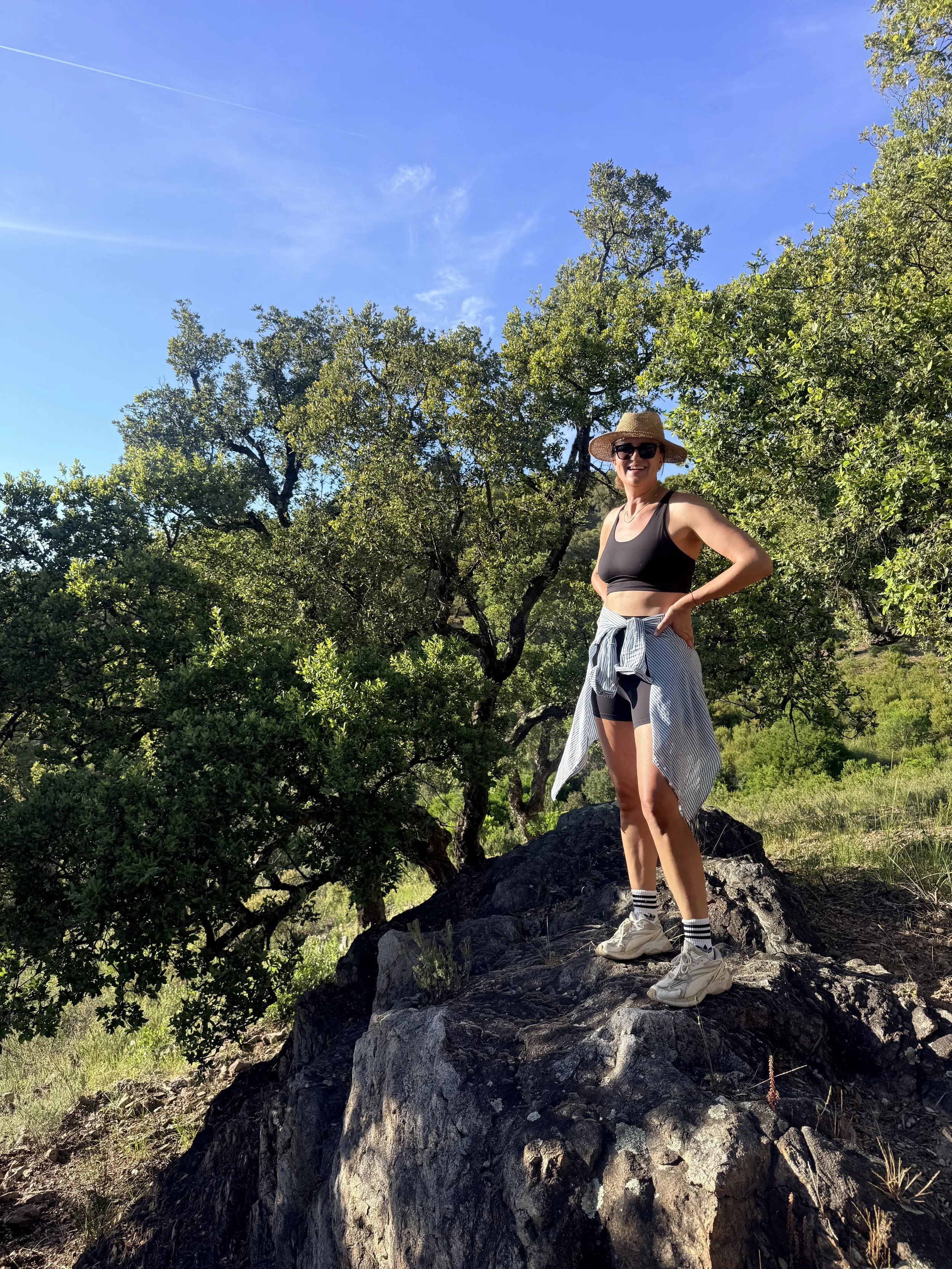 A woman standing on a large rock outdoors, wearing a straw hat, sunglasses, a black tank top, and striped shorts, with a background of green trees and a blue sky.