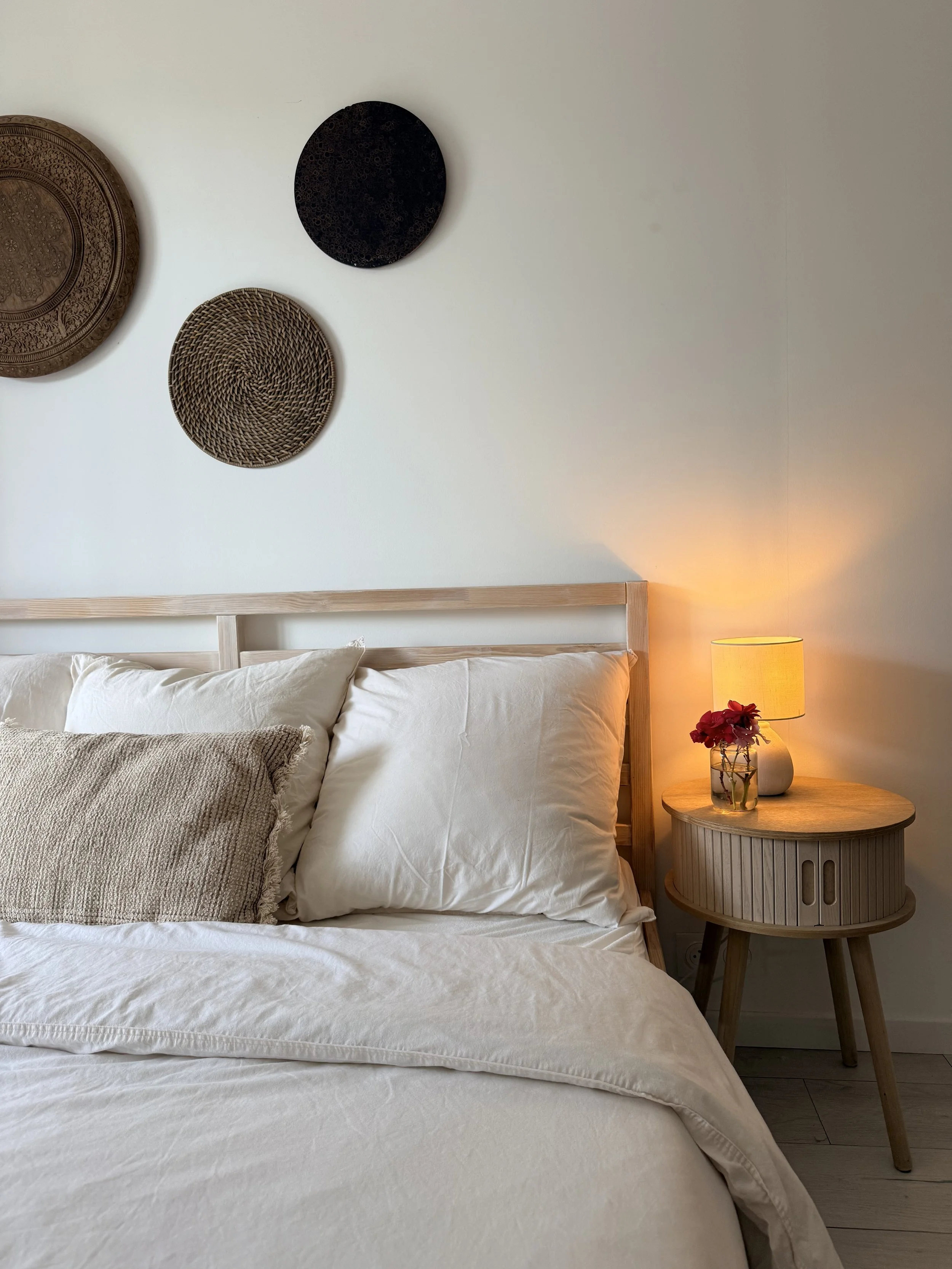 A bedroom with a wooden bed frame, white bedding, multiple pillows, a small bedside table, a lamp, and wall decor including three decorative plates.