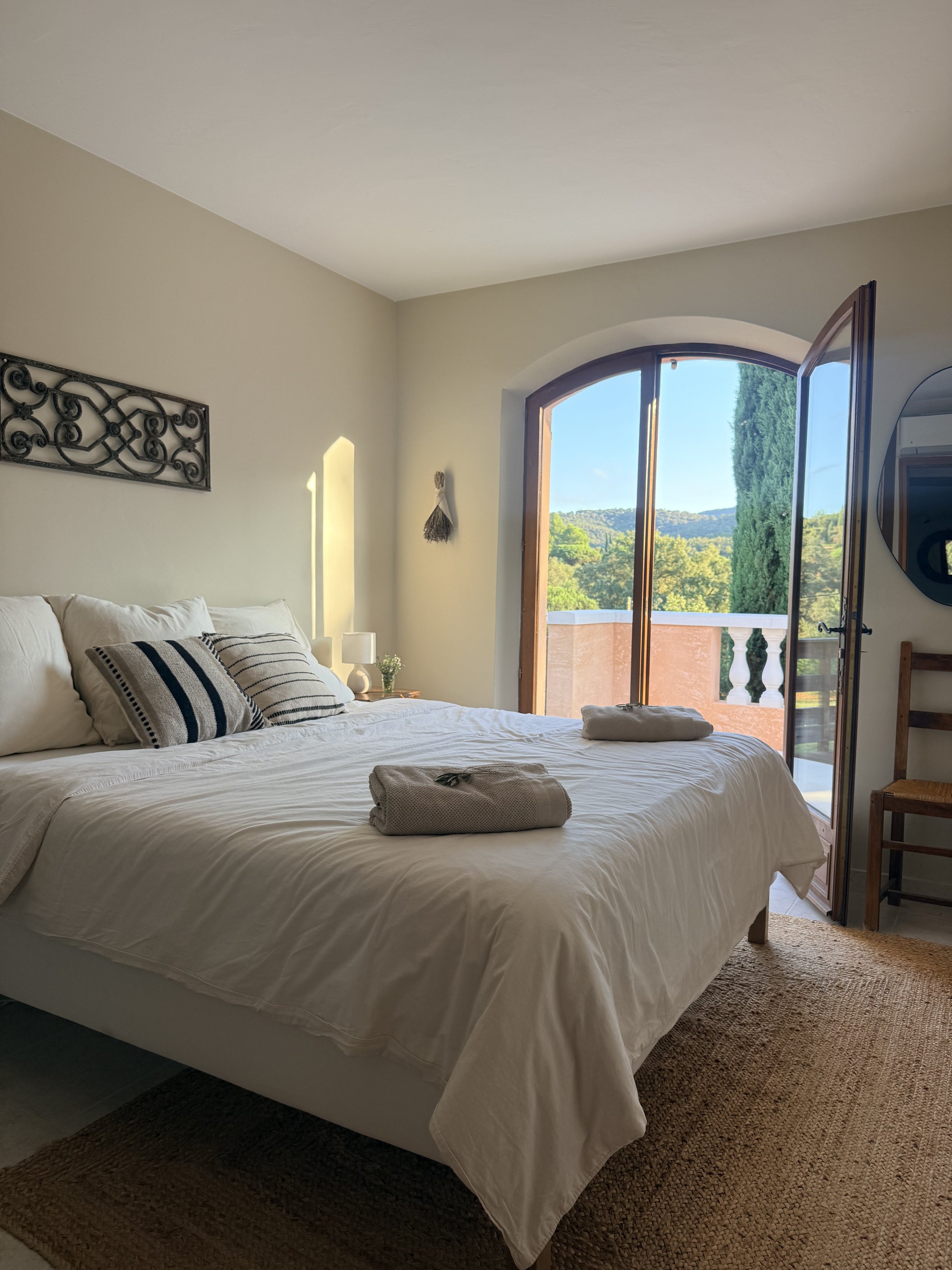 Bedroom with white bedding, pillows, open door leading to balcony with mountains and trees outside, sunlight streaming in, mirror on wall, wicker rug on floor.
