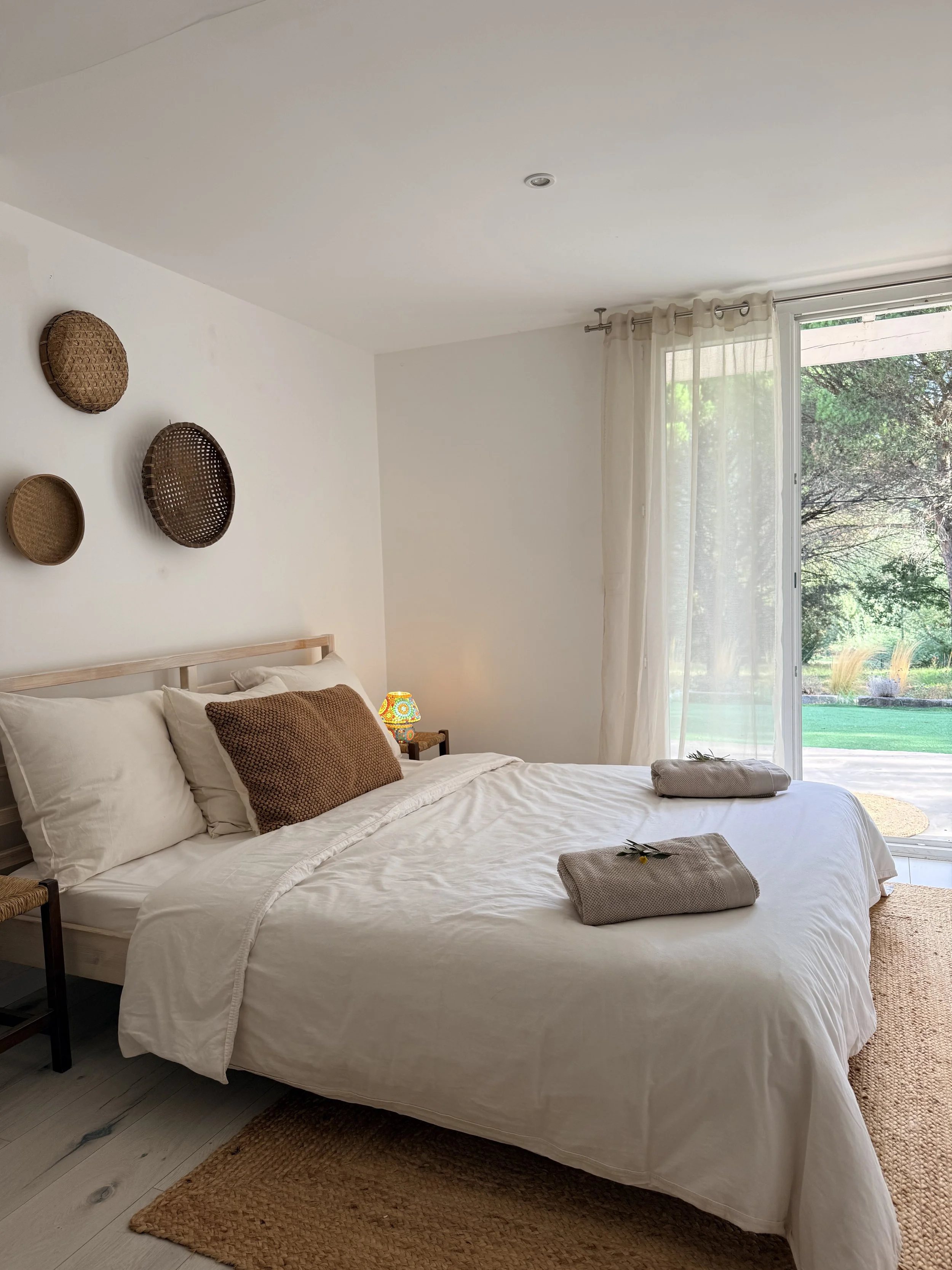 A bedroom with a bed covered in white bedding, two rolled towels on the bed, brown pillow, side table with a decorative lamp, large window with sheer curtains, and view of a green outdoor yard.