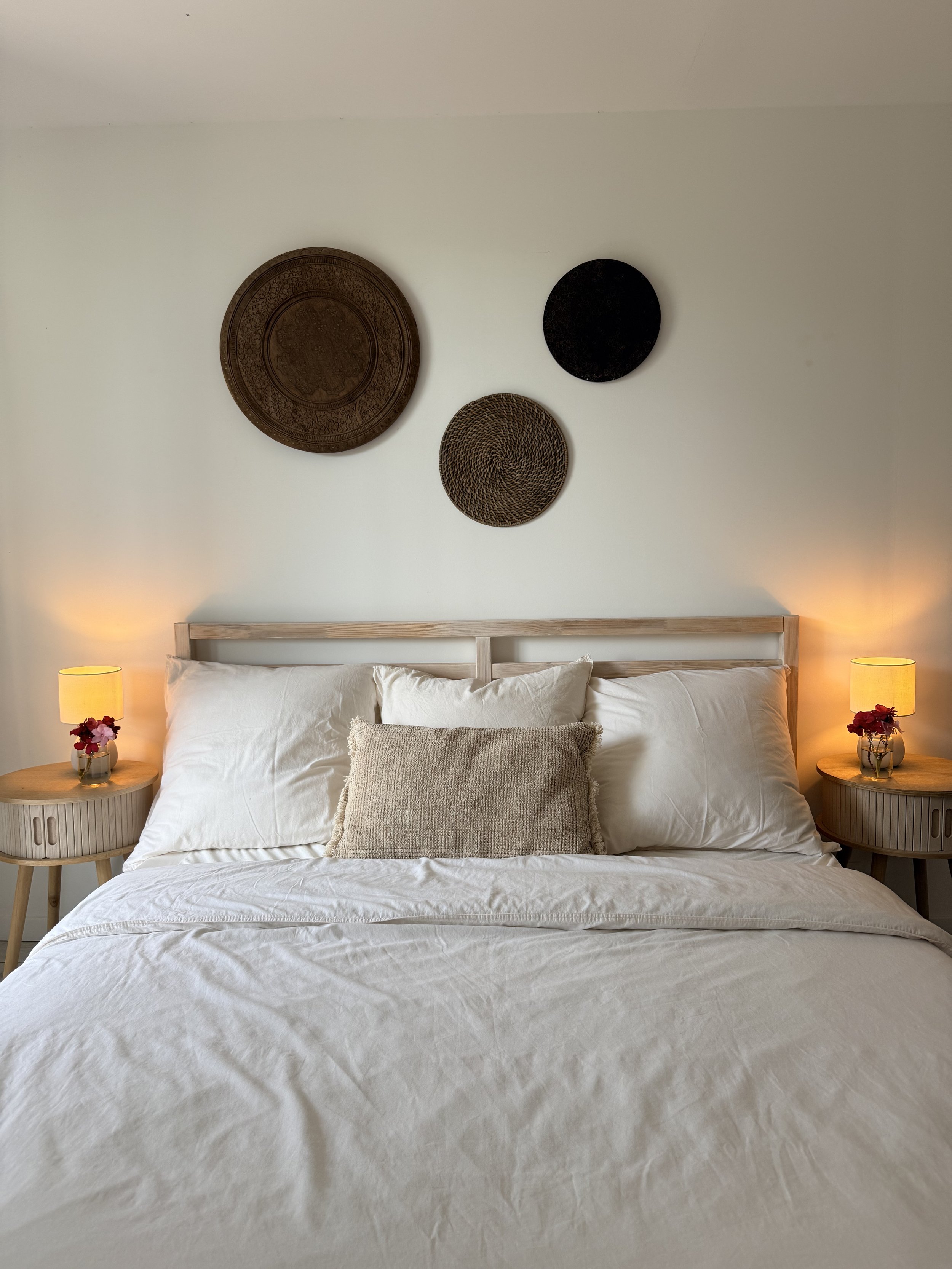 A neatly made bed with white sheets, pillows, and a beige cushion, flanked by two wooden nightstands with yellow lamps and small floral arrangements, with decorative wall art above the bed.