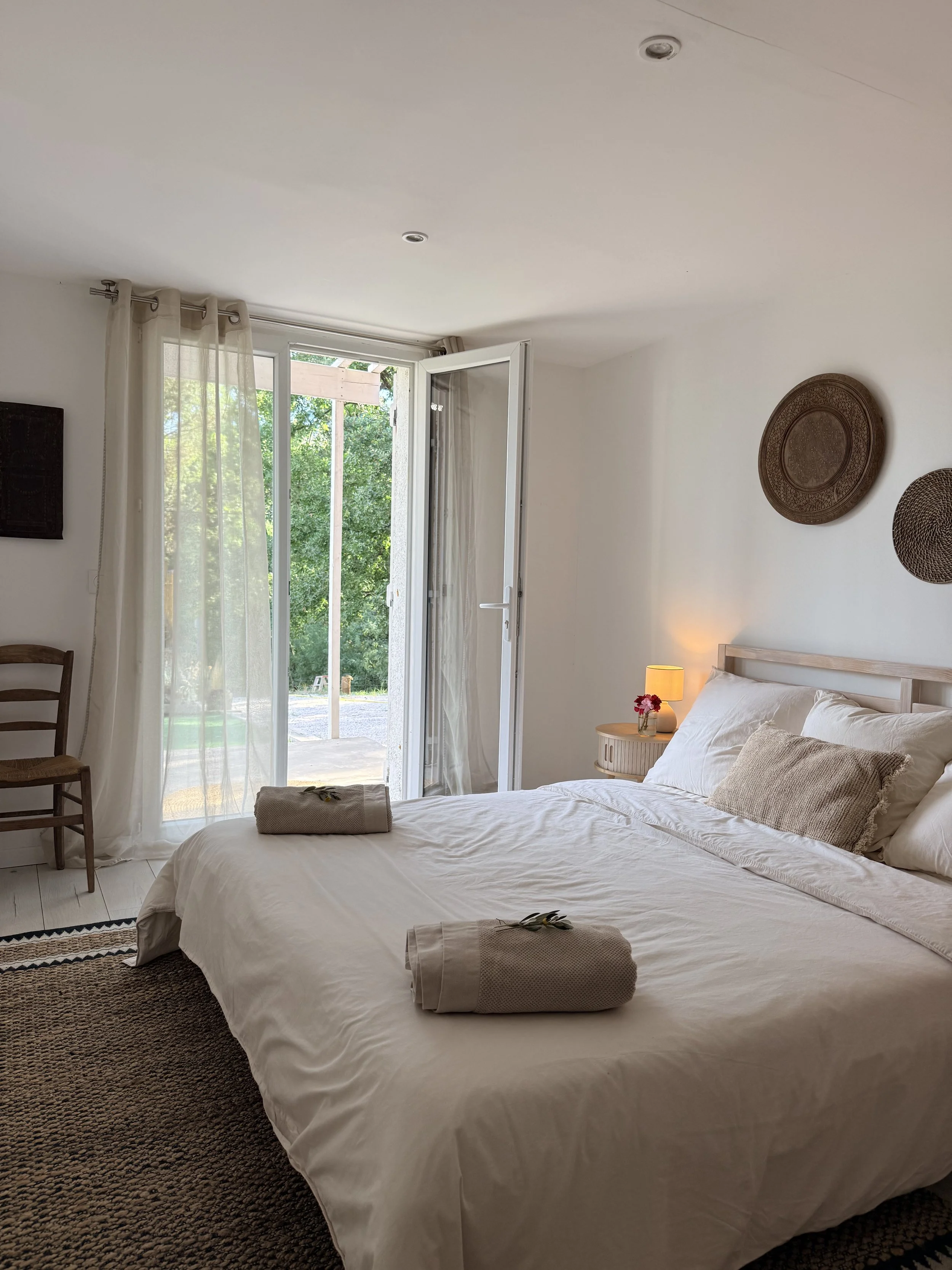 A bedroom with a neatly made bed, a small side table with a lamp and flowers, open glass doors leading outside with greenery visible, sheer curtains, a wooden chair, and decorative wall hangings.