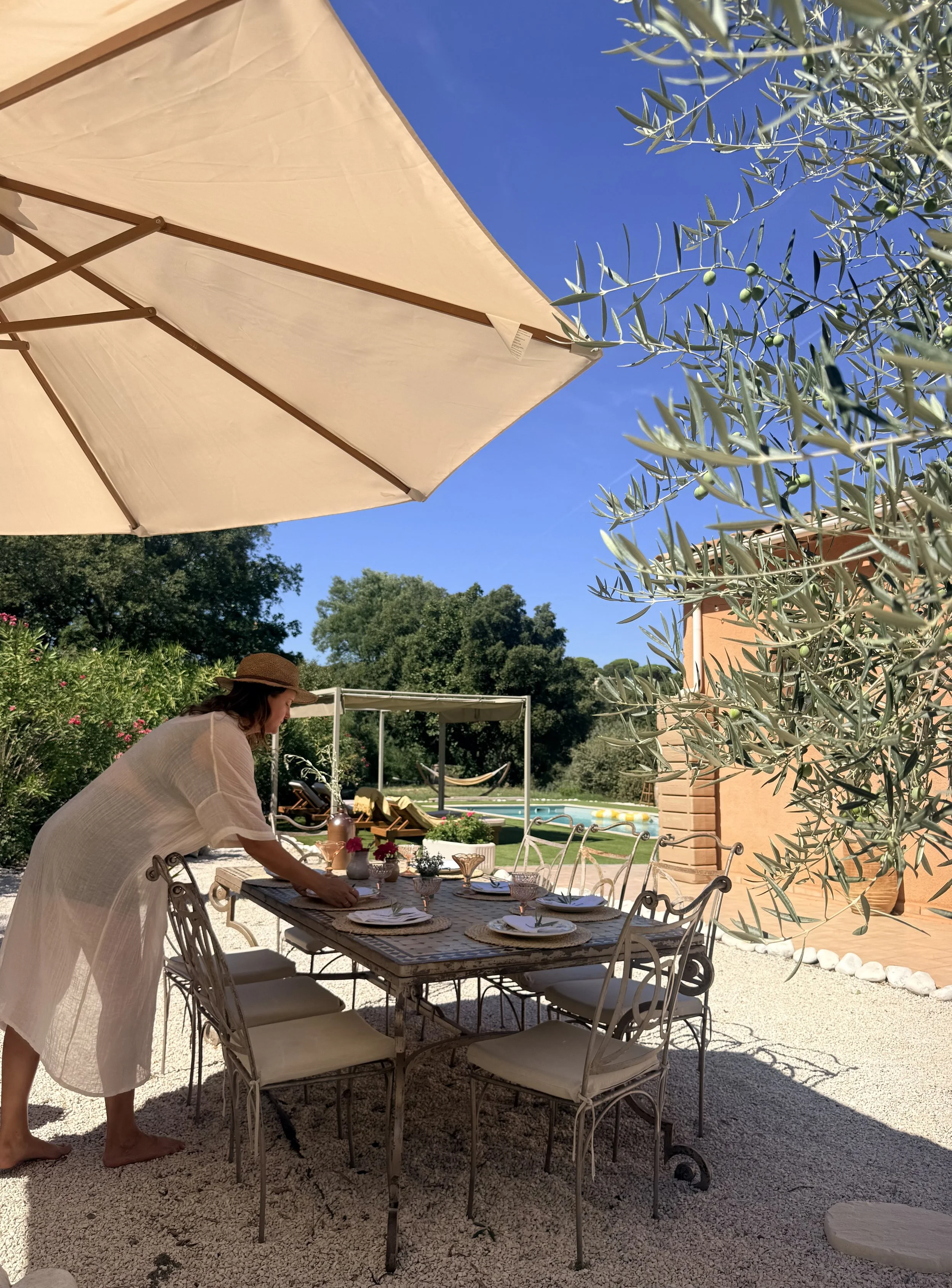 A woman prepares a table for an outdoor meal under a large beige umbrella on a sunny day. The table is set with plates, glasses, and a centerpiece. There are trees, a swimming pool, and a house in the background.