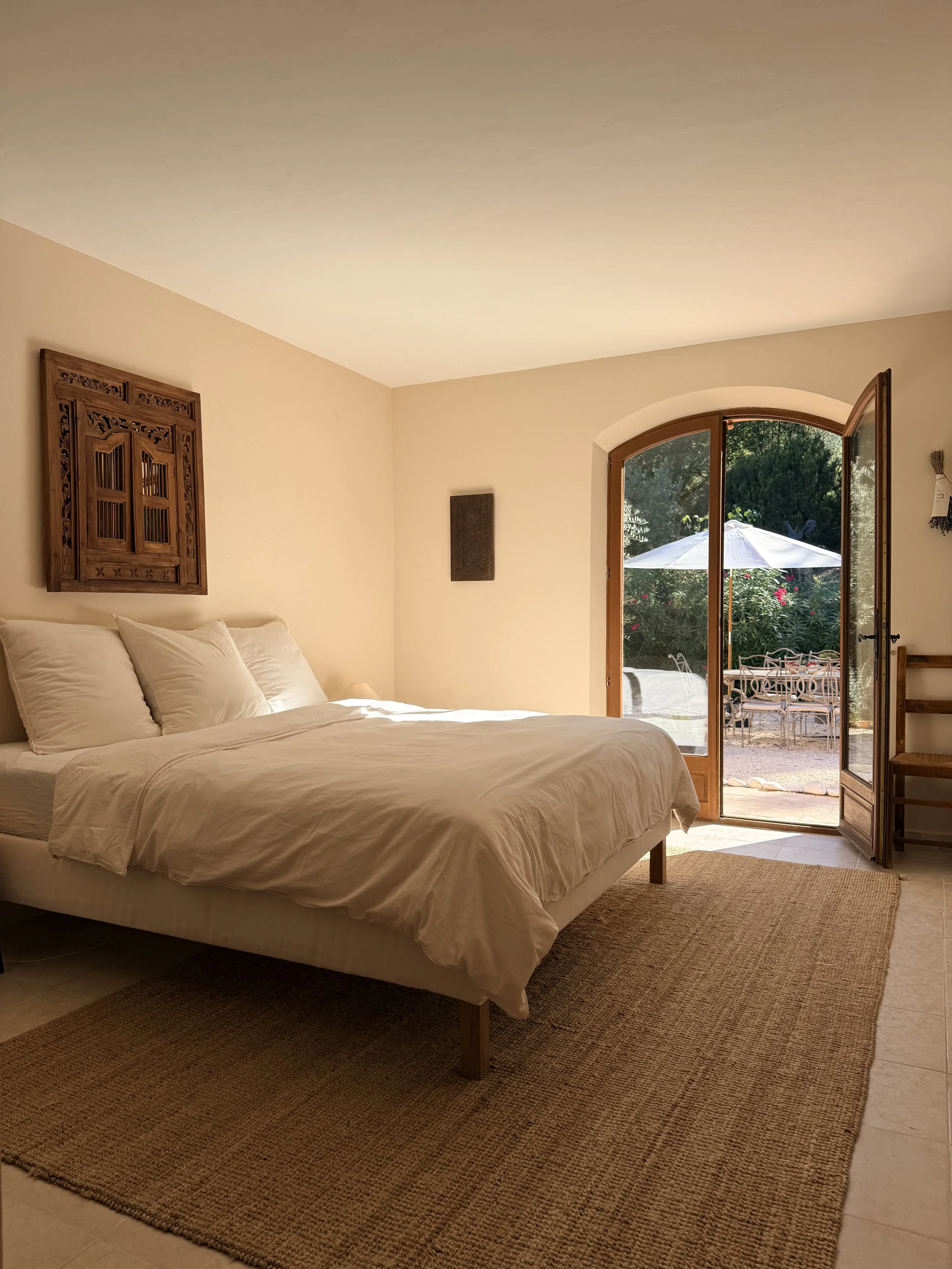A bedroom with a white bed, beige walls, and a large arched window open to a patio with outdoor furniture and greenery.