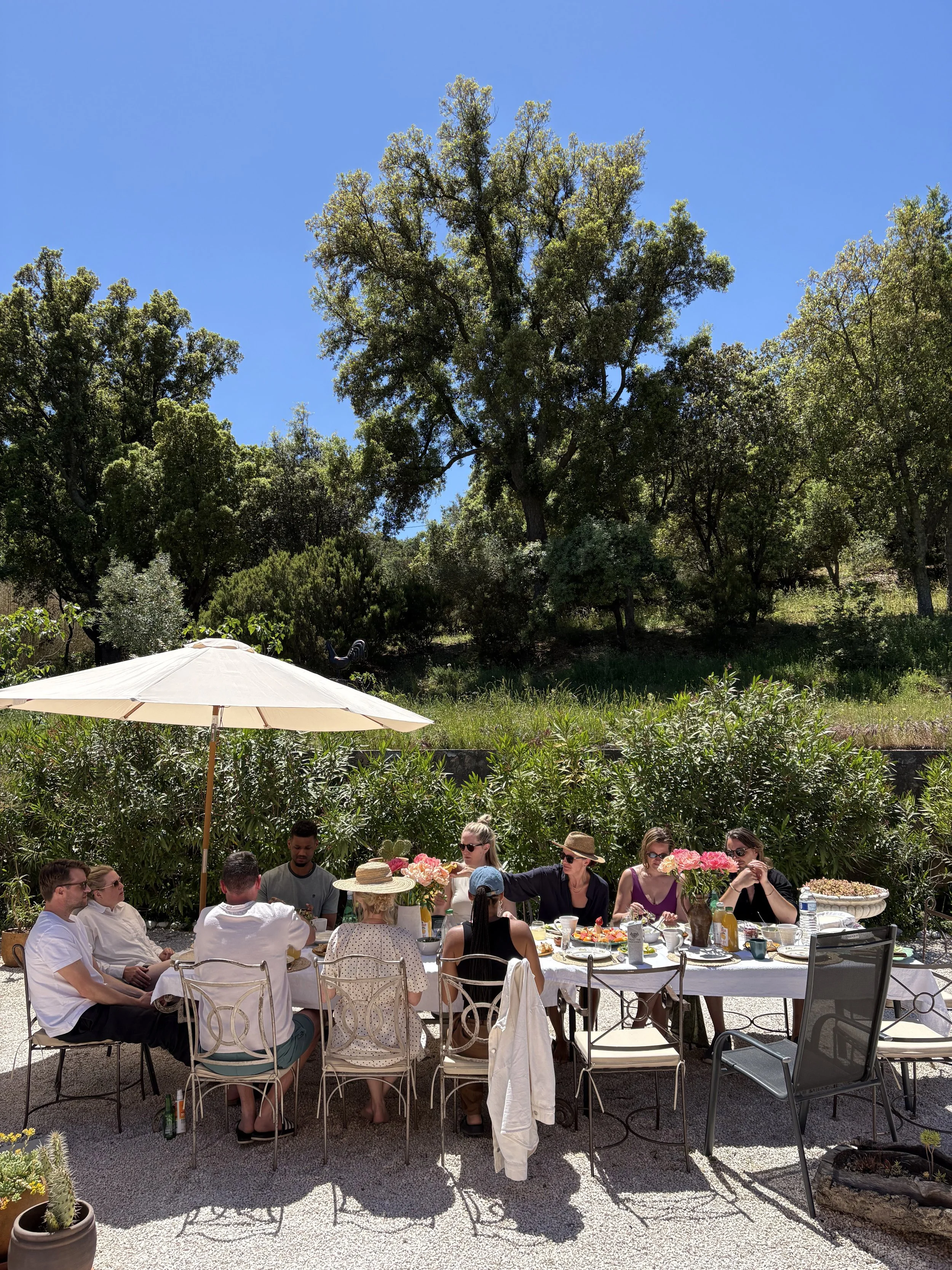 People having a meal outdoors under a large white umbrella with trees and blue sky in the background.