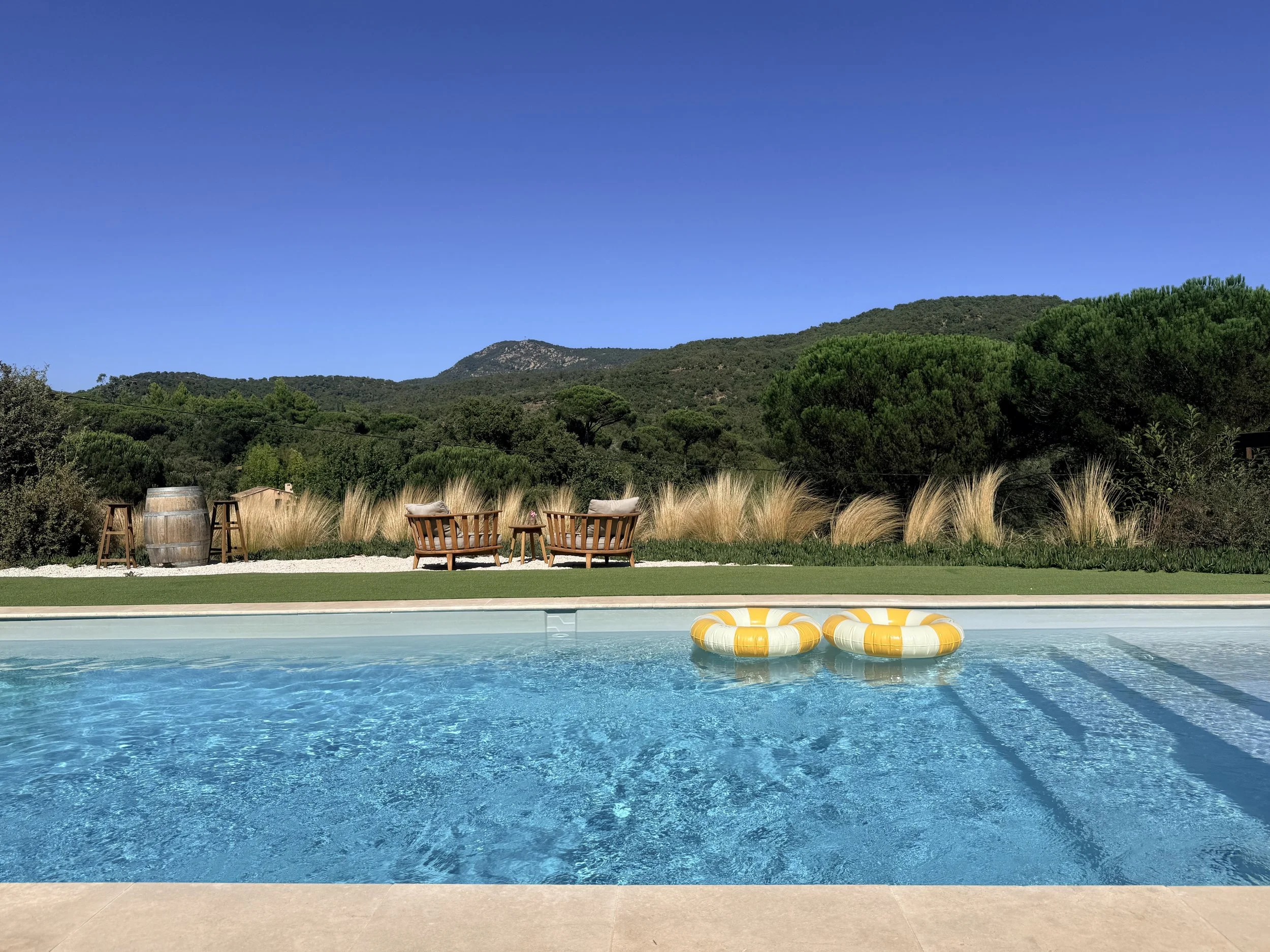 A swimming pool with clear blue water, floating life rings, and surrounding chairs on the patio. In the background, there are green trees and mountainous hills under a clear blue sky.