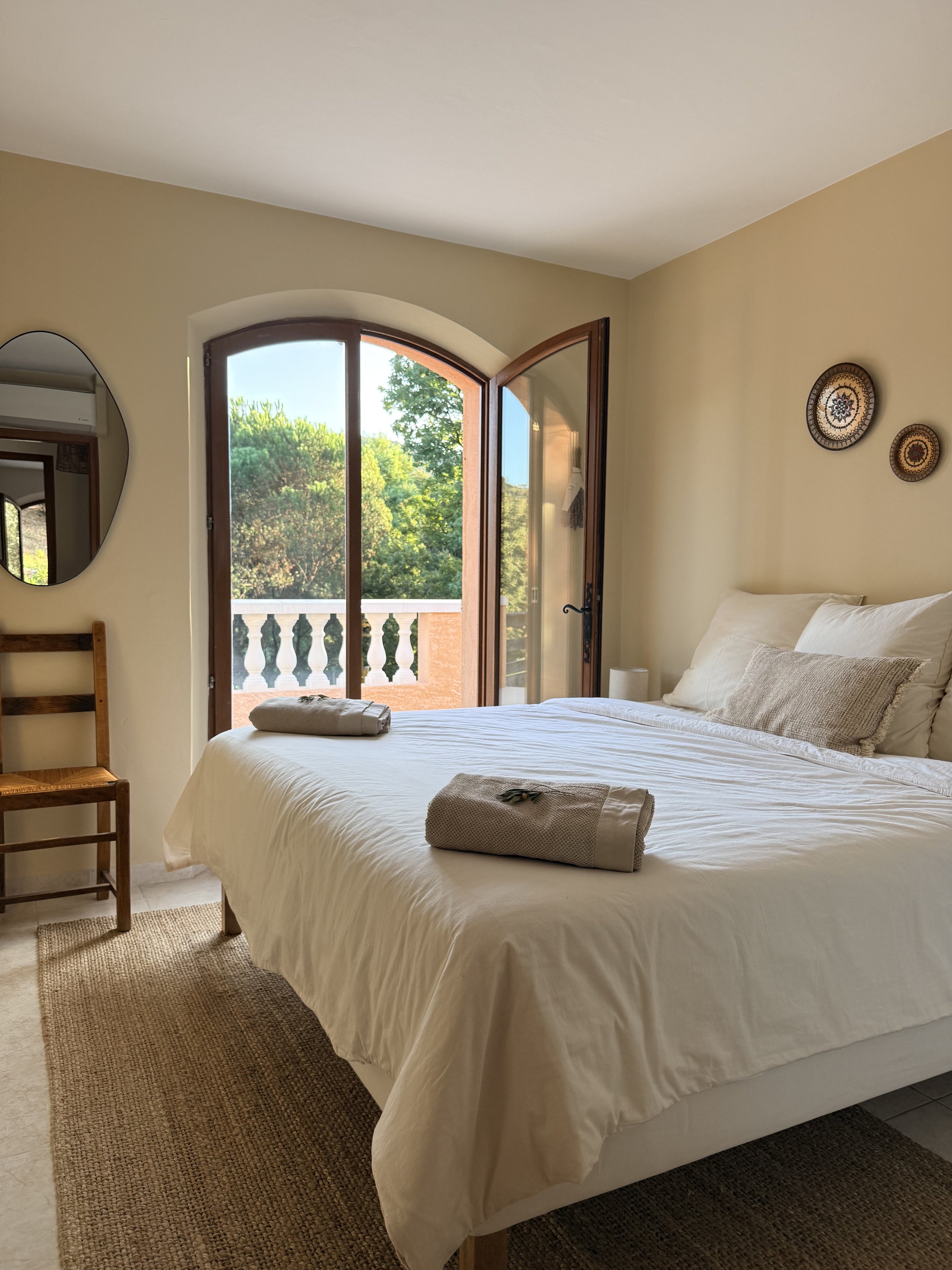 A bedroom with a large bed covered in white bedding. Towels are placed at the foot of the bed. There is a wooden chair on the left, a mirror on the wall, and decorative plates on the wall. The room has a large arched window with a view of trees and a