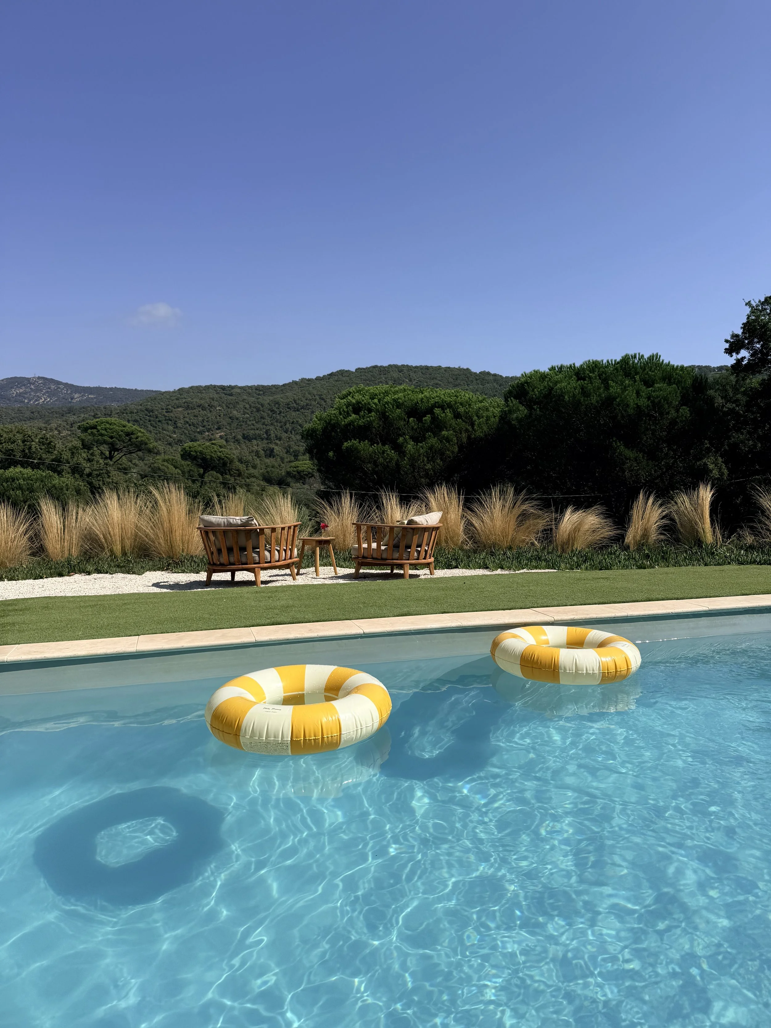 A swimming pool with yellow and white lifebuoys floating on the water, surrounded by a grassy area with trees and hills in the background under a clear blue sky.