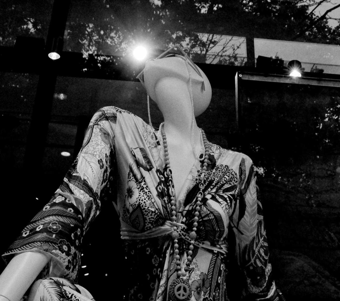 Bir mağaza podyumu üzerinde, takı takmış bir kadın mannequin'i. Model, desenli uzun kollu elbise ve büyük boncuklu kolye takıyor. Arka planda cam pencereden dış mekân görünümü.