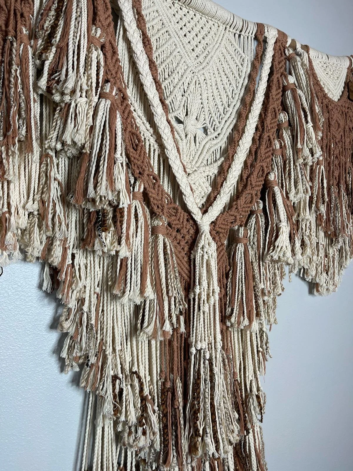 A wall hanging made of woven and knotted yarn in white and brown colors with fringe details.