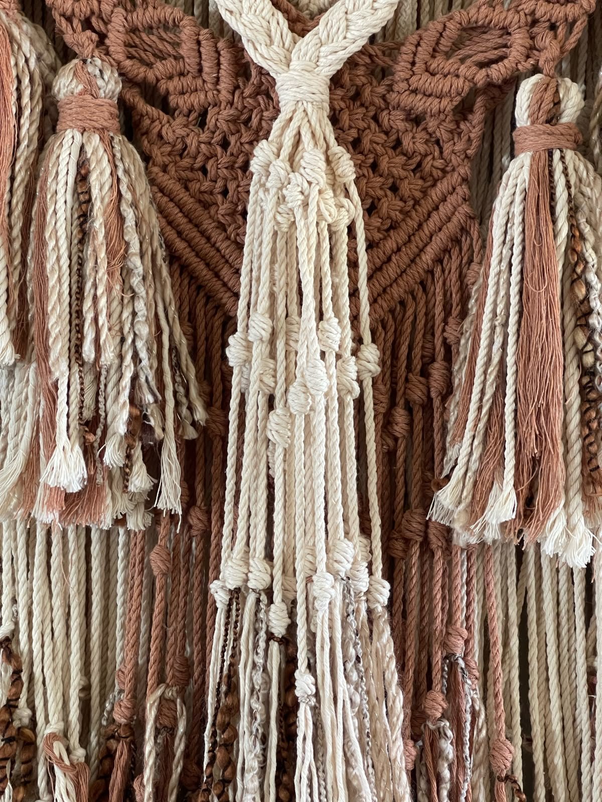 Close-up of a woven wall tapestry made of ropes in shades of brown, beige, and white with tassels hanging from the bottom.