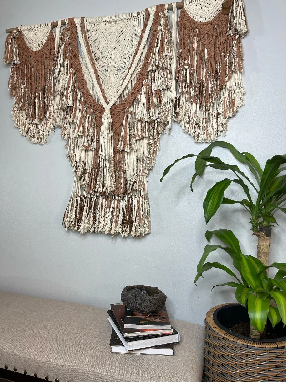 A beige upholstered bench with a stack of books and magazines, a decorative stone, a potted plant with large green leaves, and a wall hanging made of cream and brown macrame in boho style.
