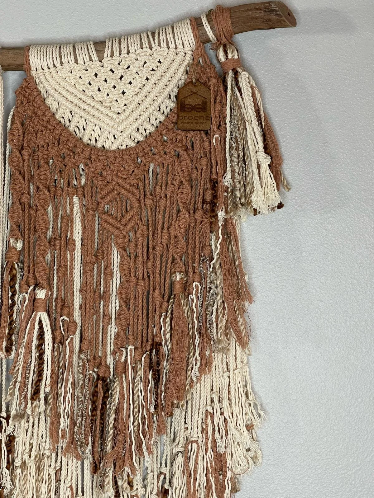 A woven wall hanging with various strands of yarn in beige, brown, and cream colors, featuring intricate textures and patterns, hanging on a light gray wall.
