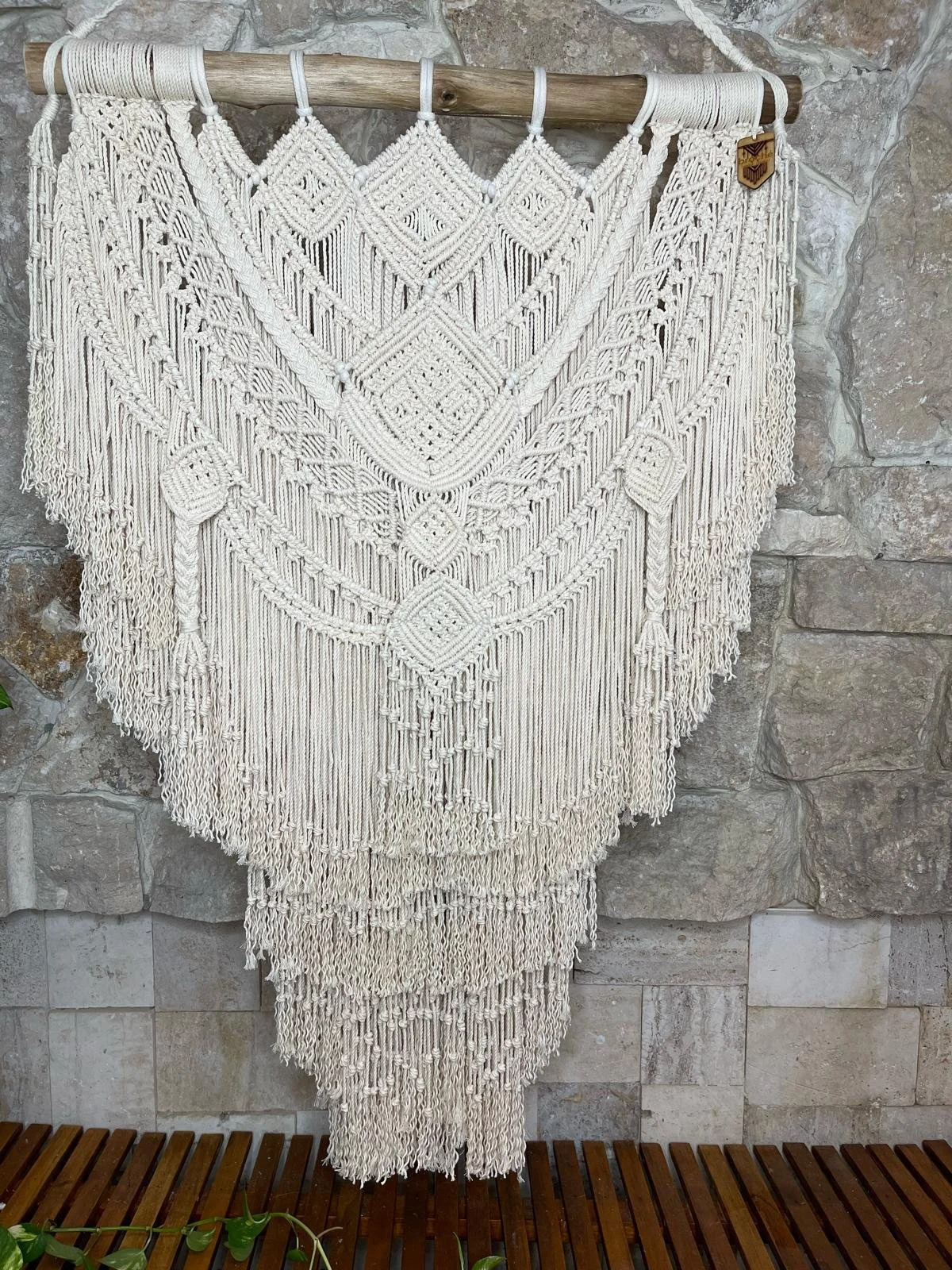 A decorative wall hanging made of white macrame with intricate patterns, hanging on a wooden rod against a stone wall.
