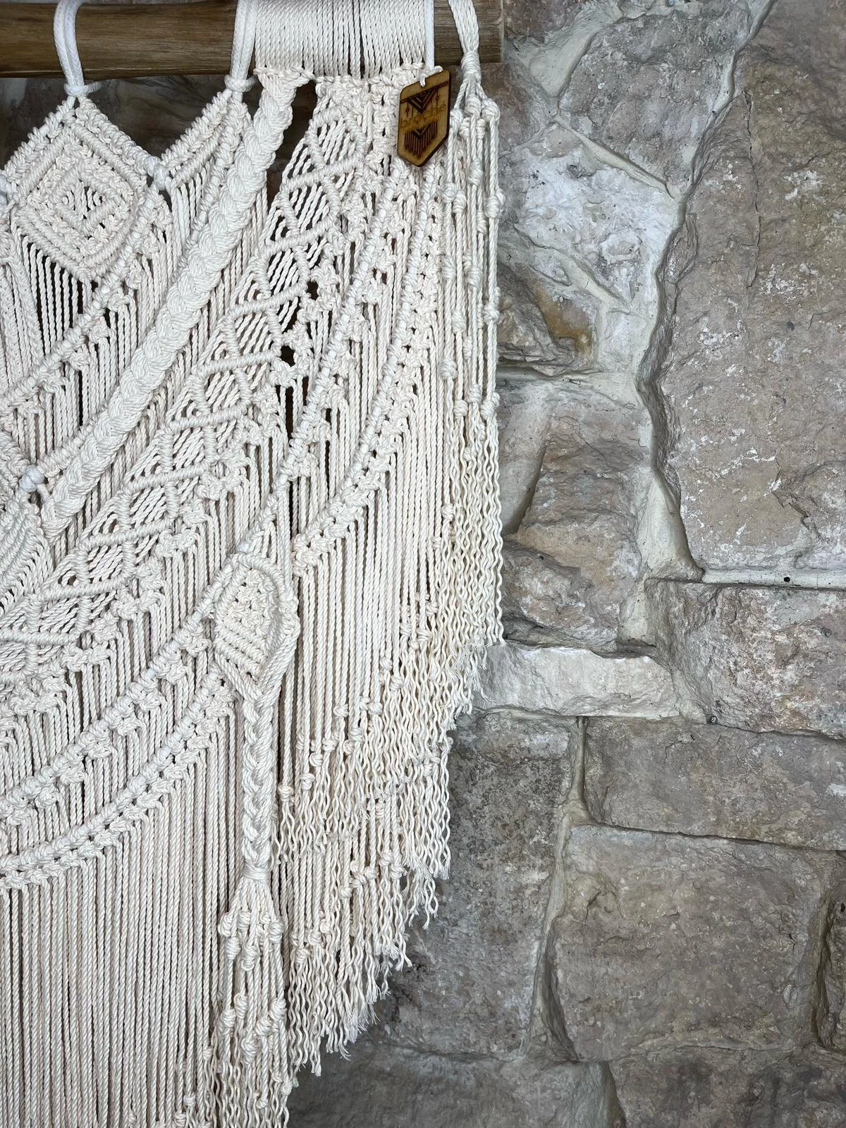 Close-up of a handmade white macramé wall hanging with intricate knots and patterns, hanging next to a rustic stone wall.