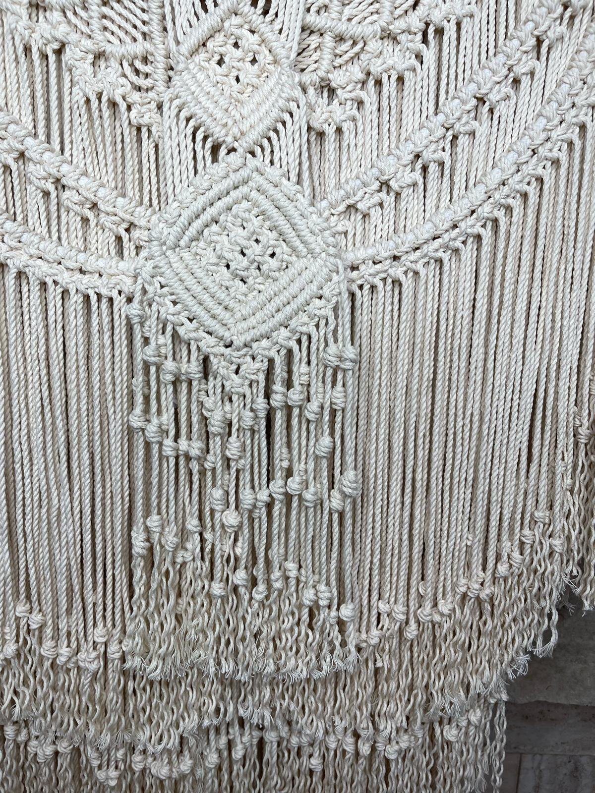 Close-up of a woven wall hanging with intricate knots and fringe details, made of white or cream-colored yarn.