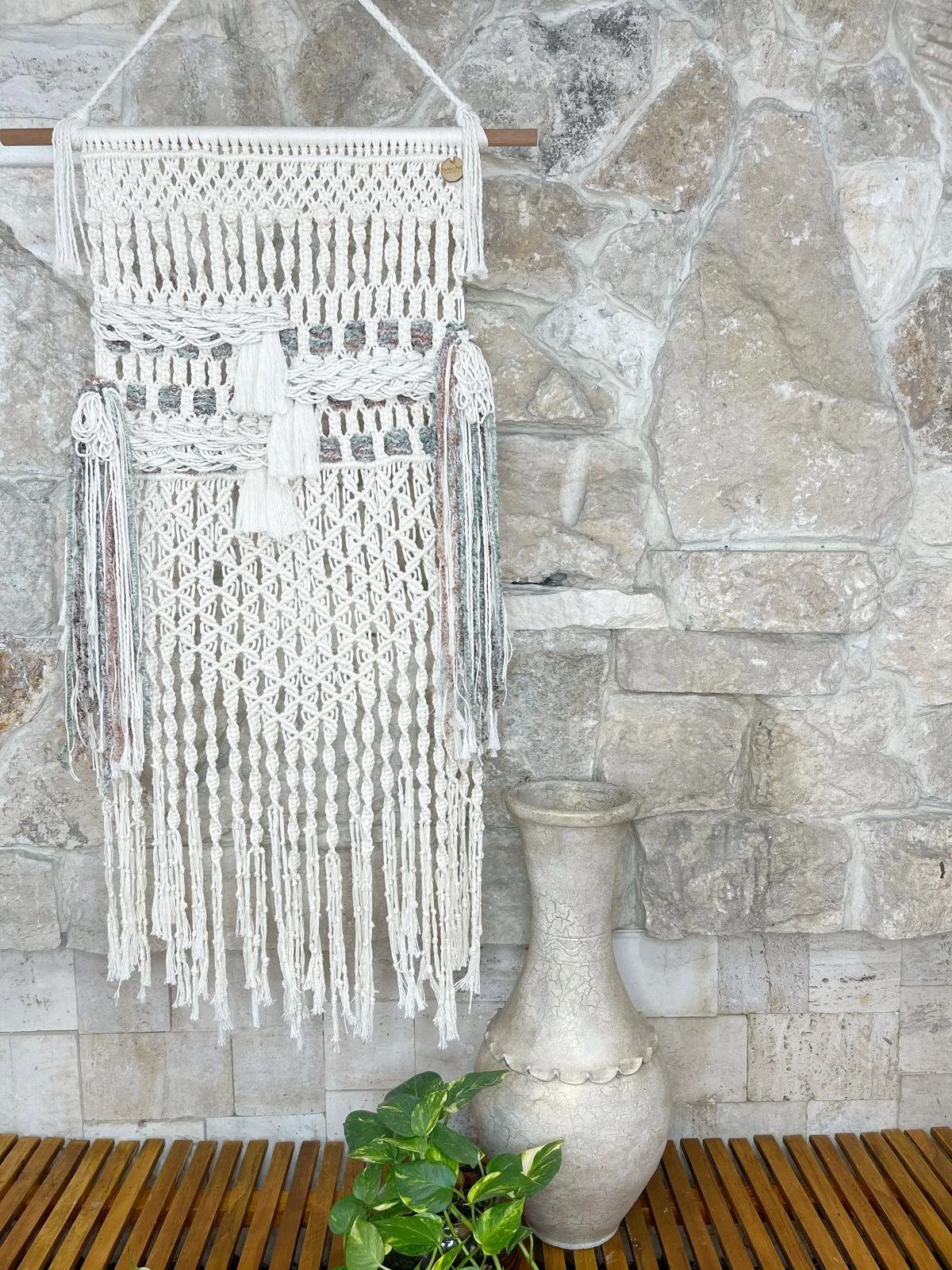 Decorative macrame wall hanging on a stone wall, with a cream-colored textured vase and potted green plant below.