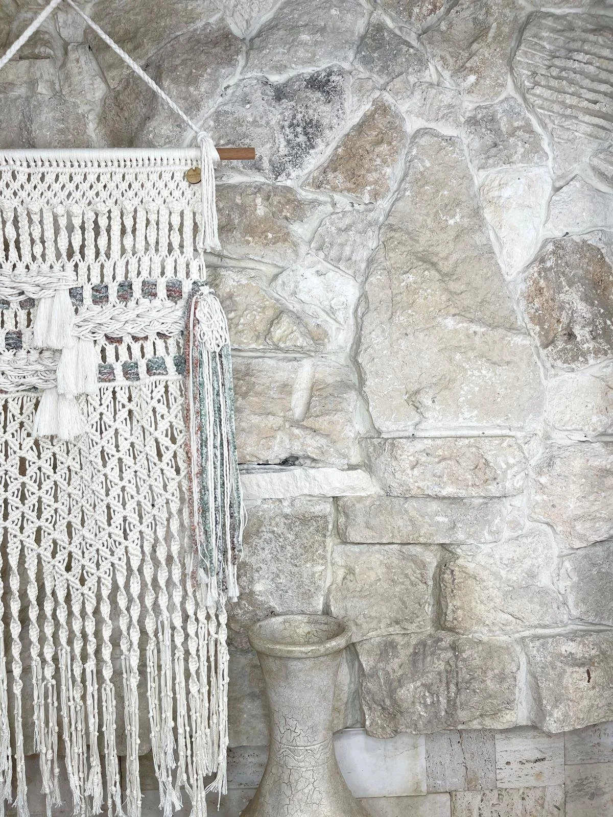 A stone wall background with a rustic, beige-colored flowerpot and a macramé wall hanging with tassels hanging on the left side.