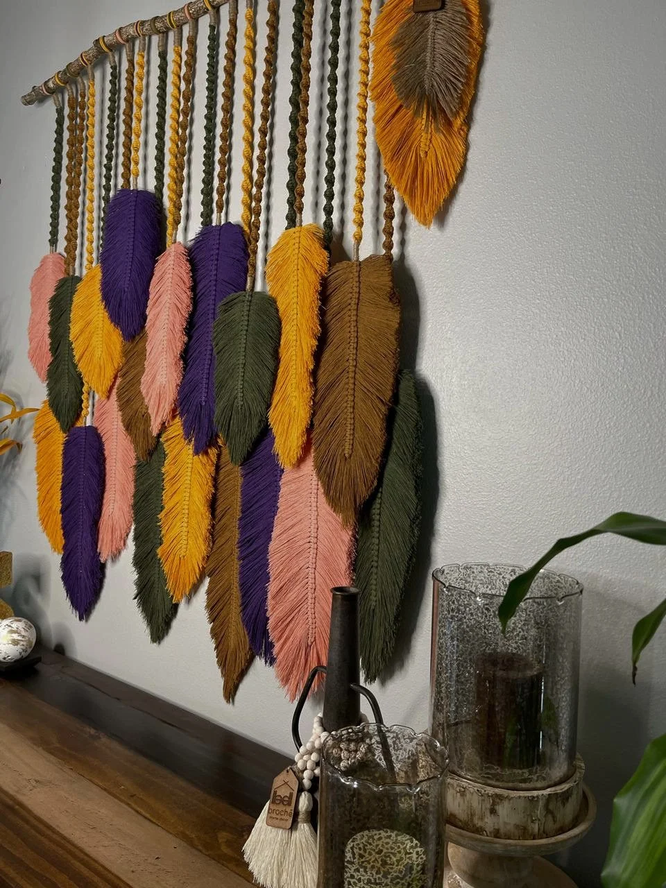 Colorful decorative wall hanging made of yarn and feathers in purple, pink, yellow, orange, green, and brown, hanging on a gray wall above a wooden table with candles and vases.