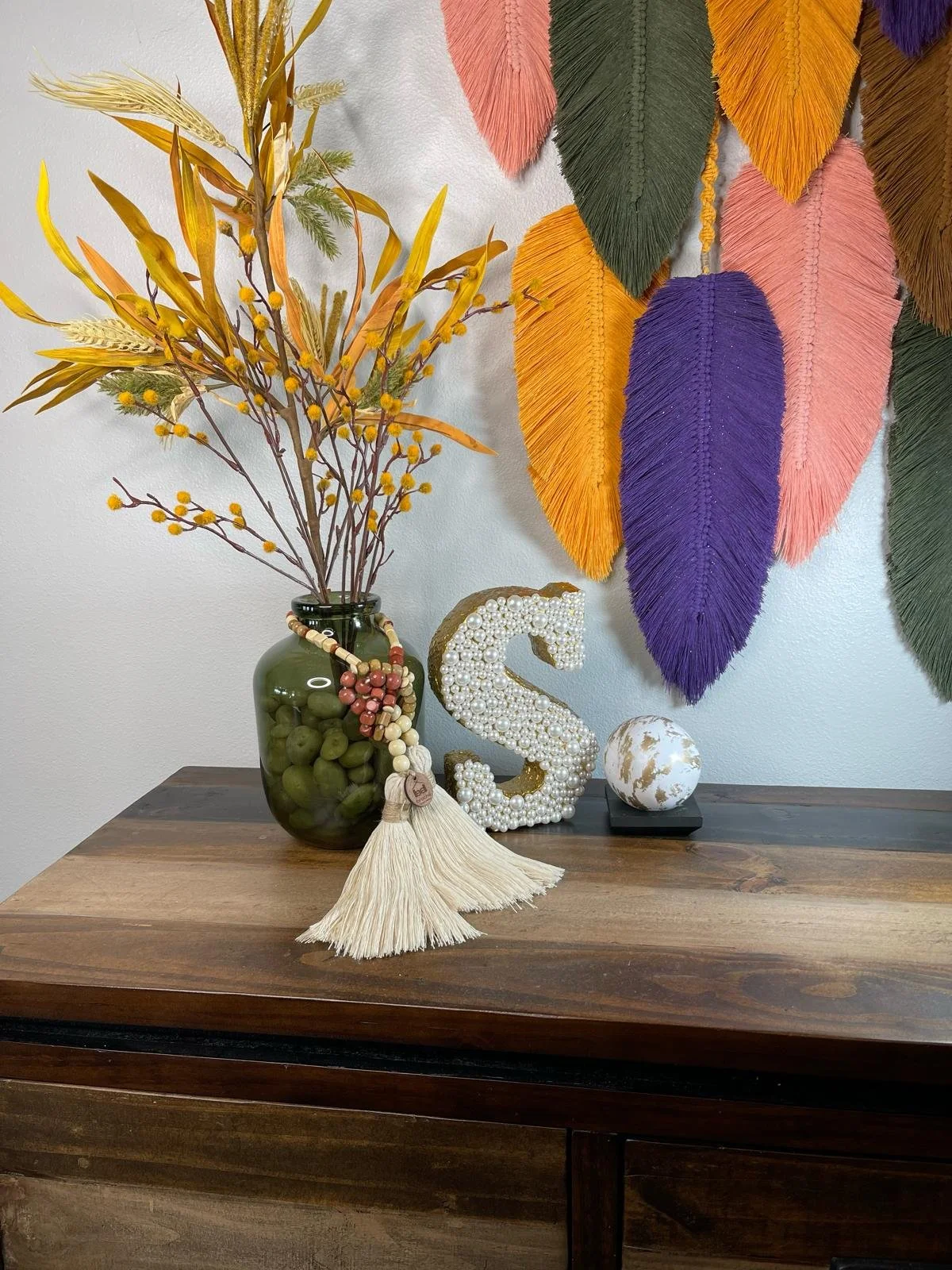 Decorative arrangement of dried flowers in a green vase, a large letter 'S' made of pearls, a white decorative ball with gold streaks, and a beaded tassel on a wooden table with colorful feather wall art in the background.