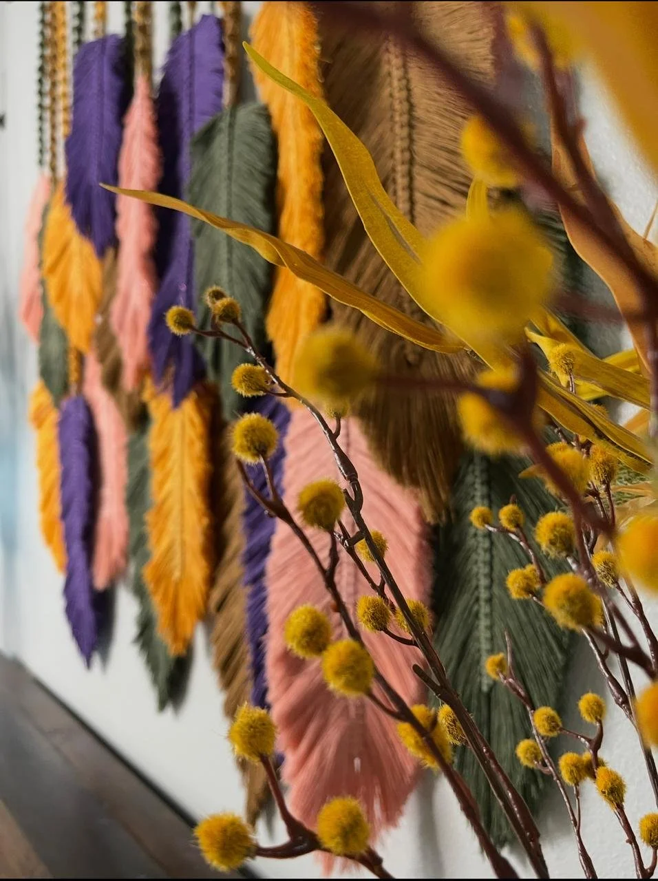 A wall decorated with colorful yarn feathers in purple, pink, yellow, and green, arranged in a pattern. In the foreground, there are yellow pom-pom branches with fuzzy spherical flowers.