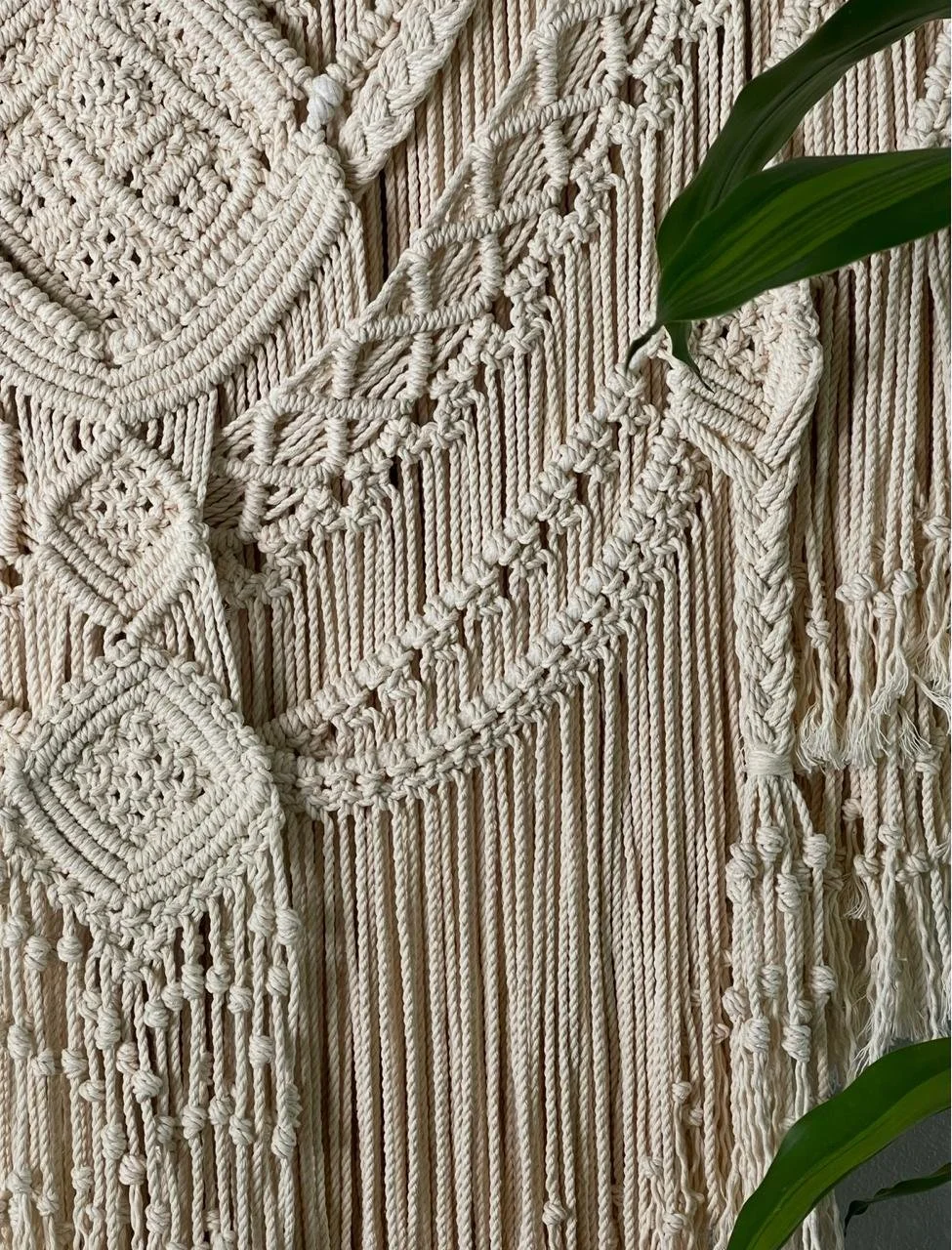 Close-up of cream-colored woven macrame wall hanging with fringe and decorative knots, partially covered by green plant leaves.