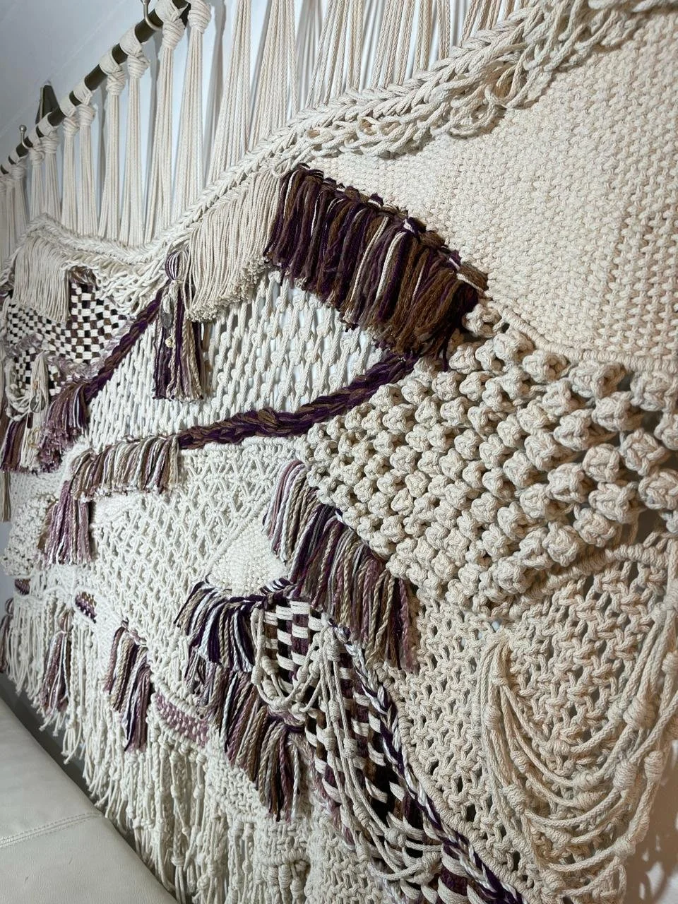 Close-up of a wall hanging decorated with various woven and knotted yarn textures and tassels in neutral colors.