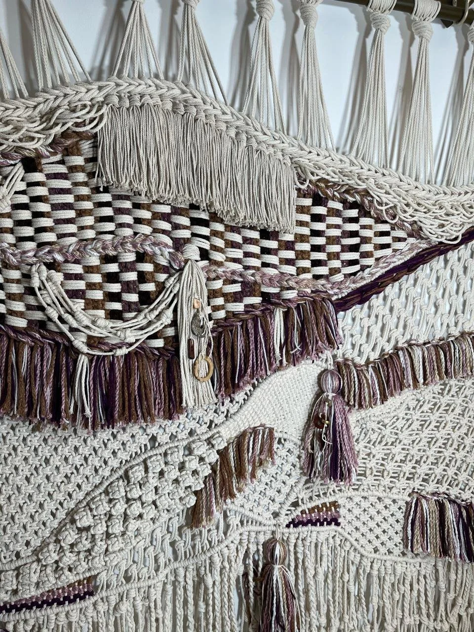 Close-up of a wall hanging with various woven and knotted textures, including tassels and fringe, in neutral tones.