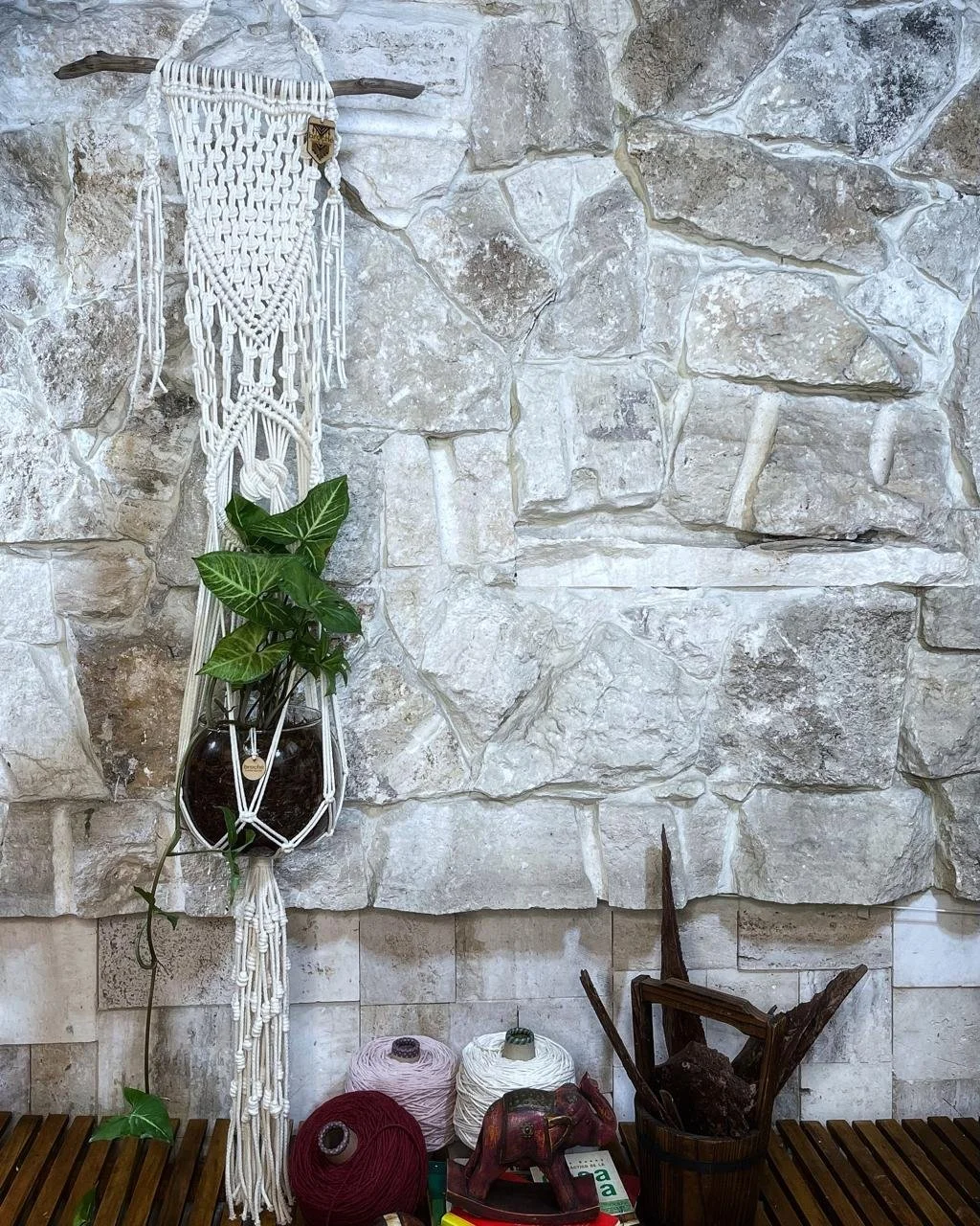 Decorative macramé wall hanging with plant in a glass container, placed against a stone wall, with several balls of yarn and woodworking tools on a wooden shelf below.