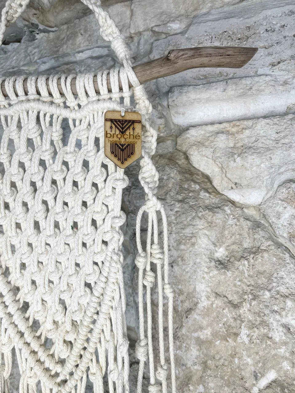 Close-up of a macrame wall hanging with a wooden branch as a support, hanging against a stone wall. The wall hanging is made of white rope with intricate knot patterns, and a wooden tag with the brand name 'Brooche' attached.