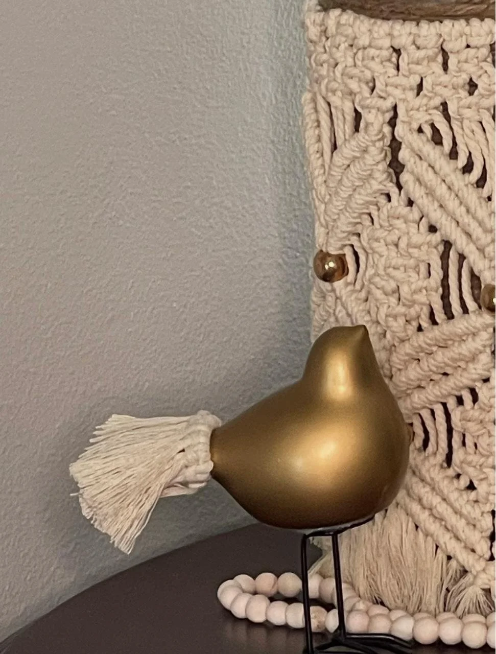 Decorative wall hanging with fabric, beads, and metallic accents, alongside a gold-colored decorative bird with a tassel tail on a black stand, placed on a dark surface with a bead garland.