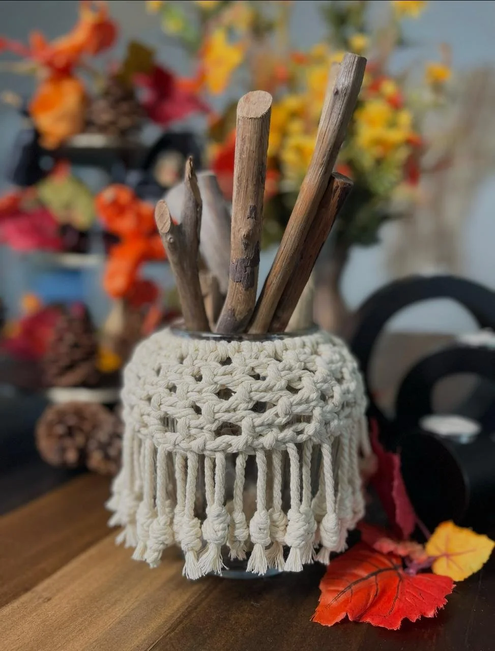 A jar with a handmade macrame cover containing wooden sticks on a wooden surface, with autumn leaves and blurred colorful fall decor in the background.