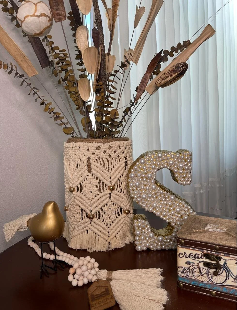 Decorative arrangement on a table includes a woven macramé-style vase with dried leaves and branches, a pearl-covered letter S, a metallic gold pear sculpture, a beaded necklace, and a small decorated box featuring a bike illustration. There are shee