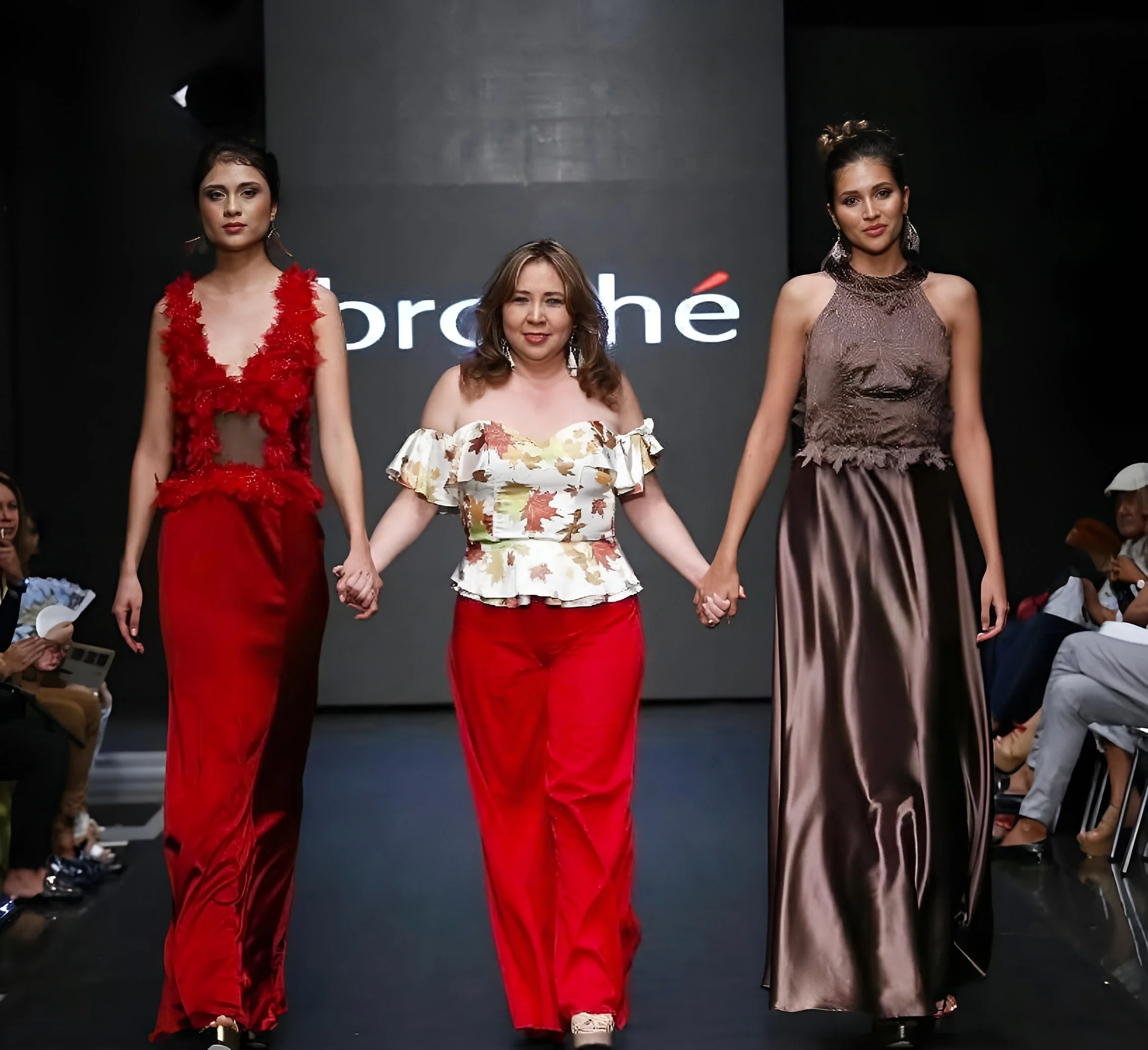 Three women holding hands on a runway during a fashion show, wearing elegant dresses in red, floral, and brown colors.