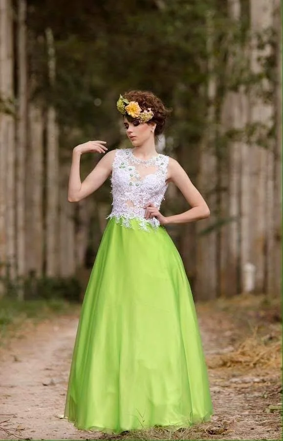 A woman in a green and white gown with floral details and a floral headpiece, standing on a dirt path in a wooded area.