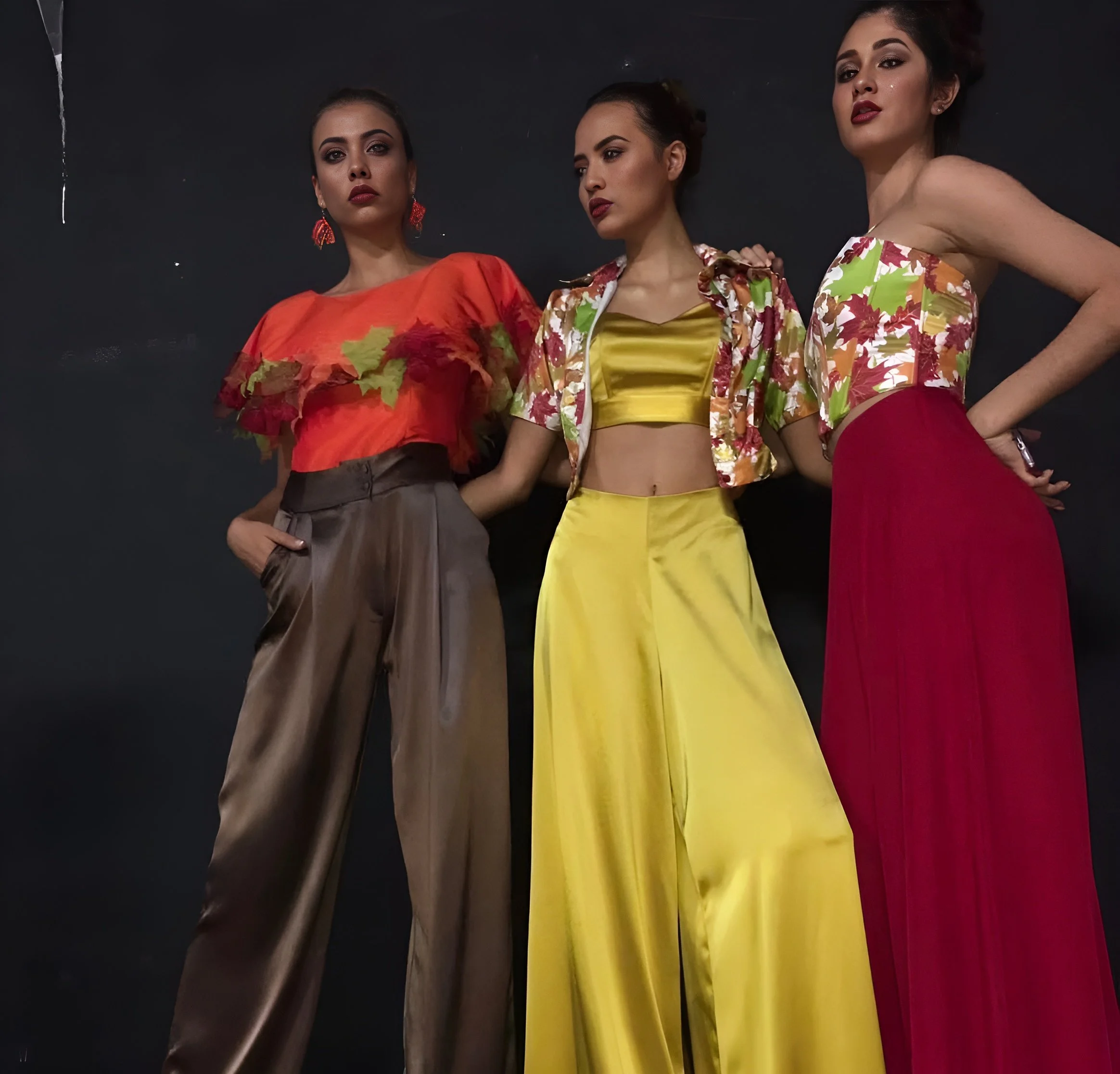 Three women dressed in colorful fashionable outfits standing against a dark background.