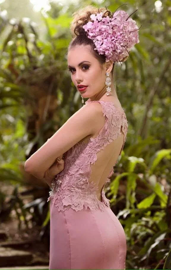 A woman in a pink lace dress with floral embellishments, wearing large earrings and a hydrangea flower headpiece, standing outdoors in a green, lush environment.