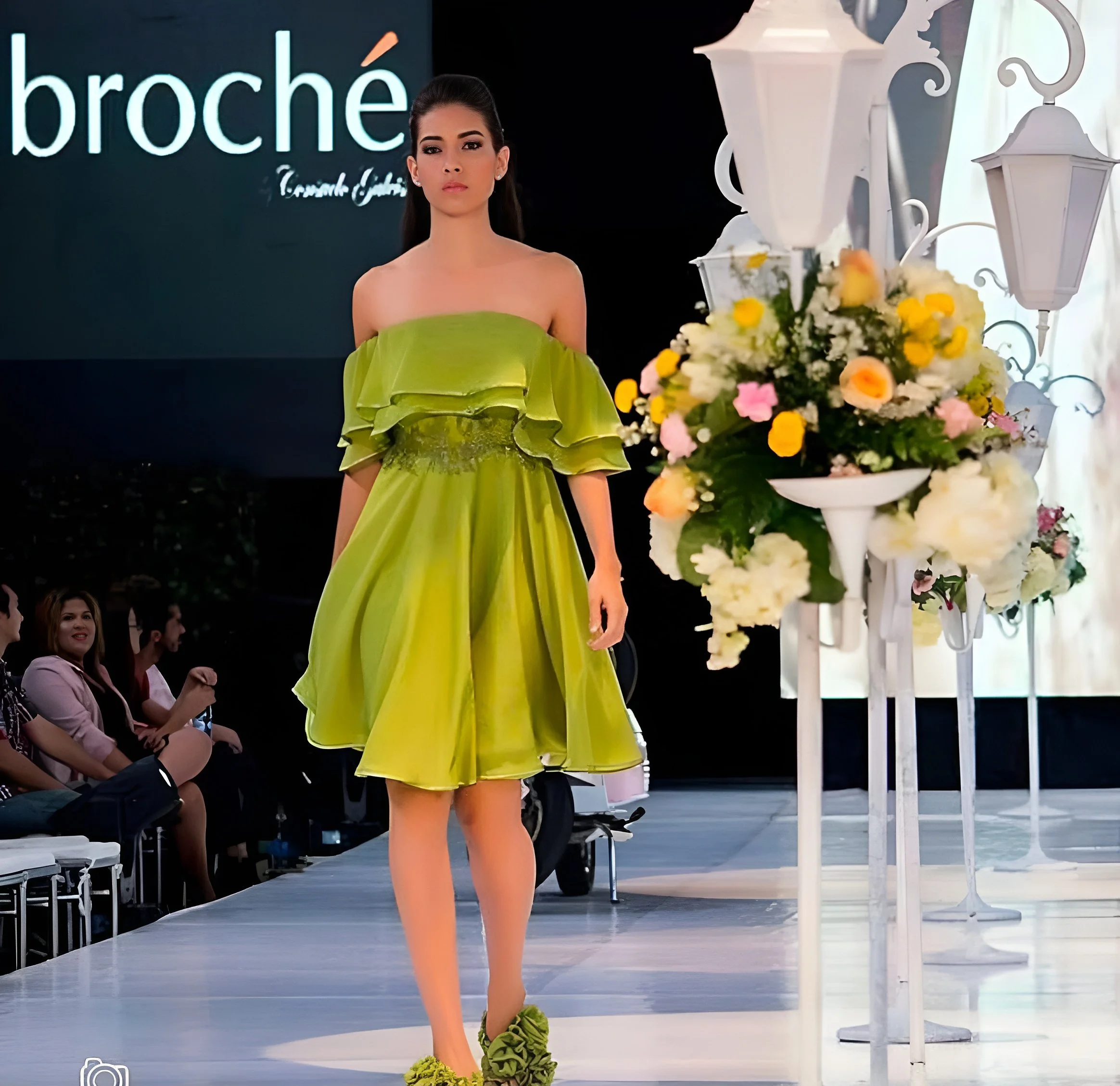 A fashion model walking down a runway during a fashion show. She is wearing a green off-the-shoulder dress with ruffle details and matching green shoes with ruffles. The background shows an audience and decorative white lanterns with flowers.