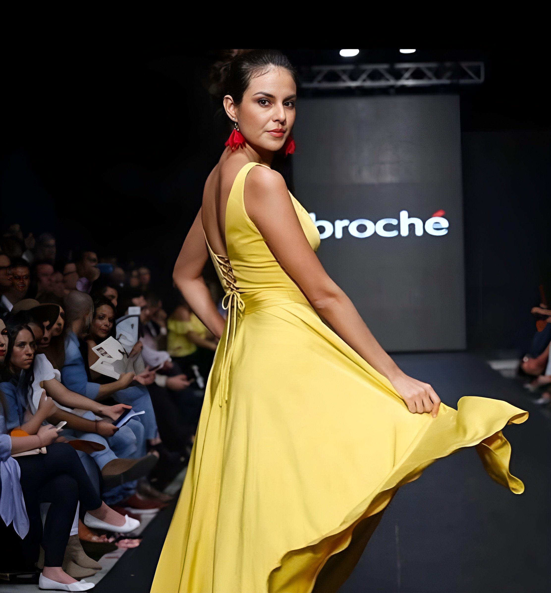 A model walking down the runway during a fashion show wearing a yellow dress with red earrings, onlookers seated on either side of the runway, with a large screen in the background displaying the brand name "Broche."