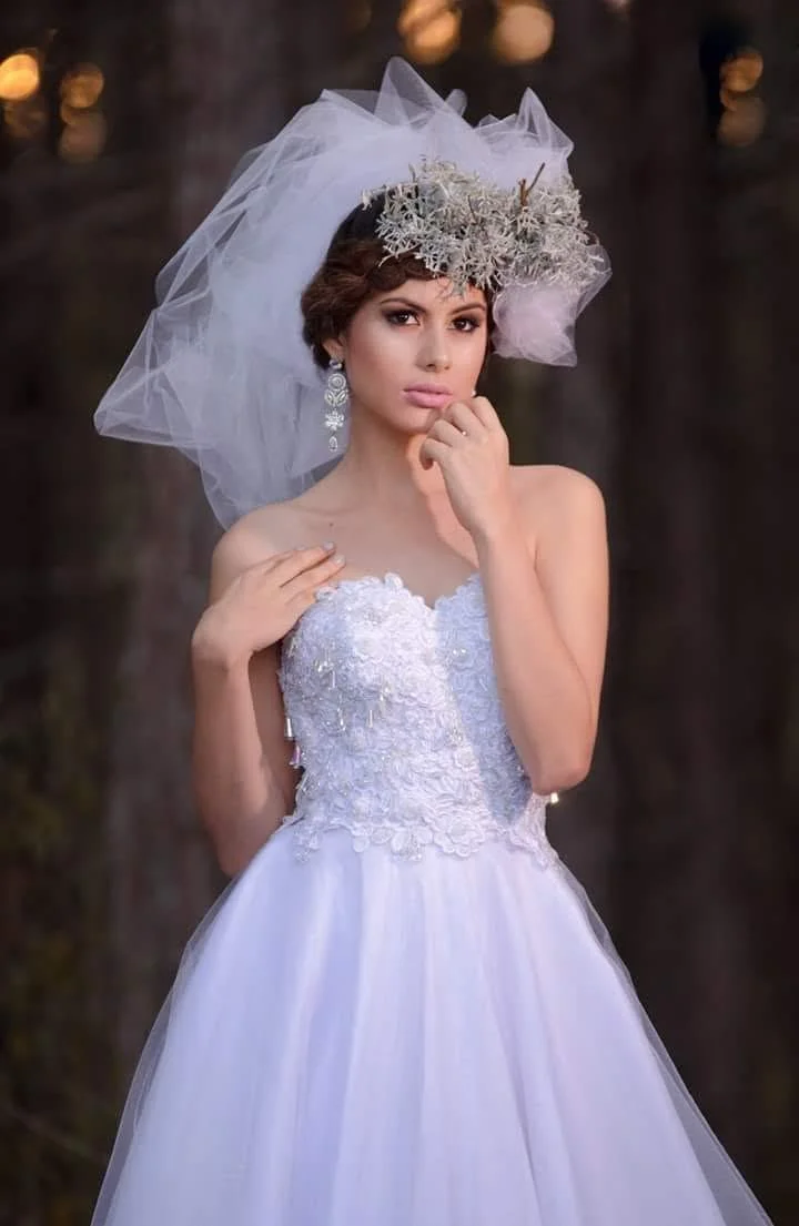 A bride in a white wedding dress with lace details, wearing a veil, and a floral headpiece, standing outdoors with blurred lights in the background.