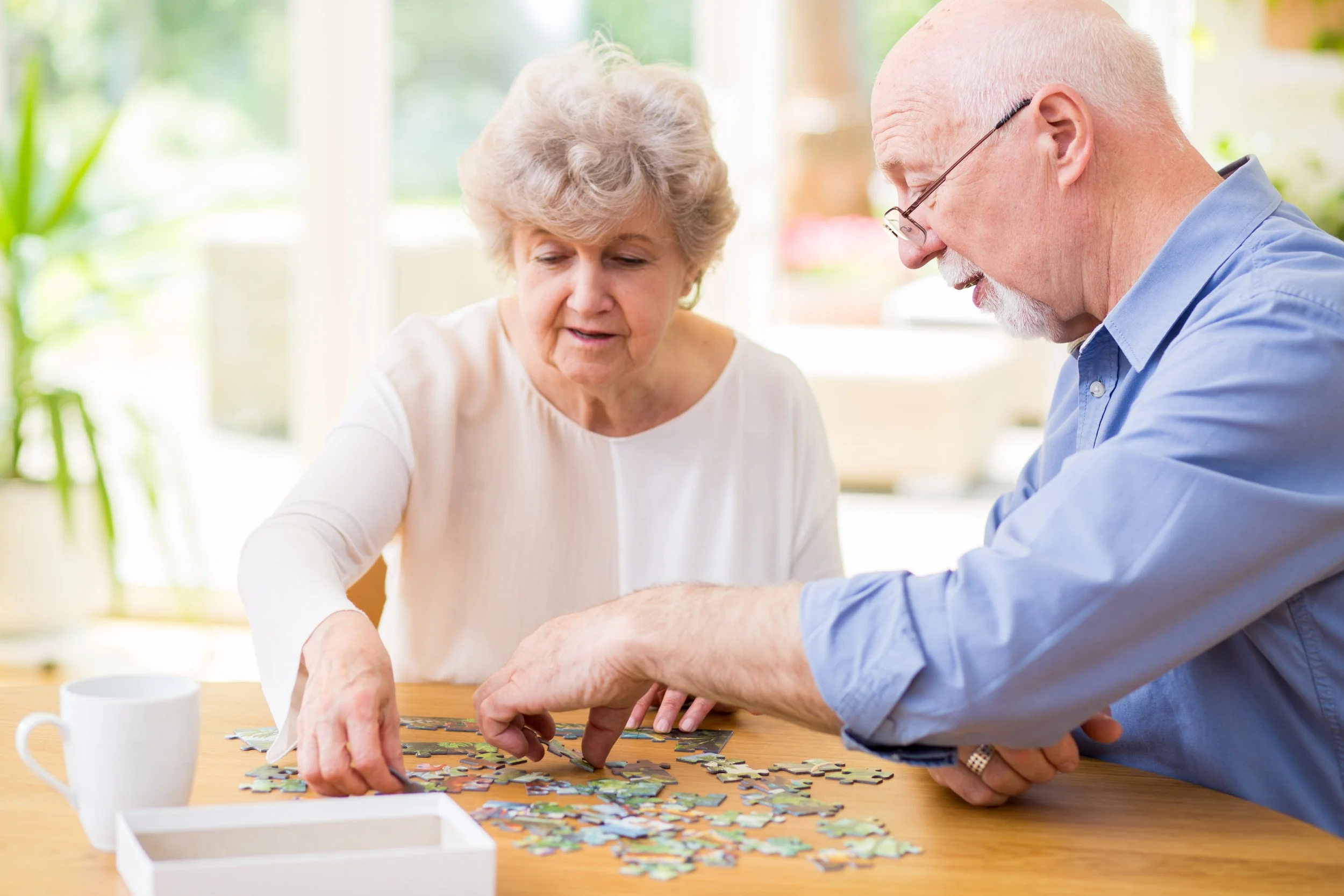 An elderly woman and an elderly man are working together on a jigsaw puzzle at a wooden table in a bright room. There is a white mug on the table and a white box in the foreground. They are focused on the puzzle, with the woman pointing and the man looking closely.